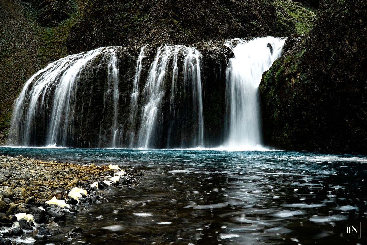A stunning wide waterfall cascades over mossy rocks into a serene turquoise pool surrounded by dark stone cliffs.