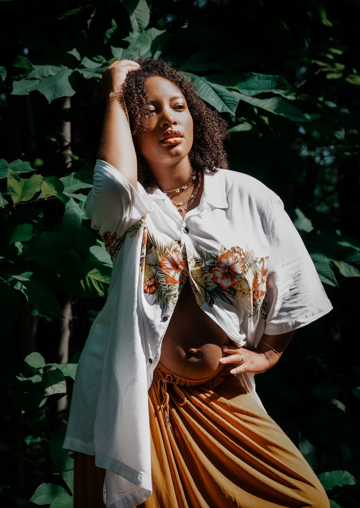 Maternity photo of a young woman in white floral crop top and gold pleated skirt posing gracefully among lush green leaves outdoors.