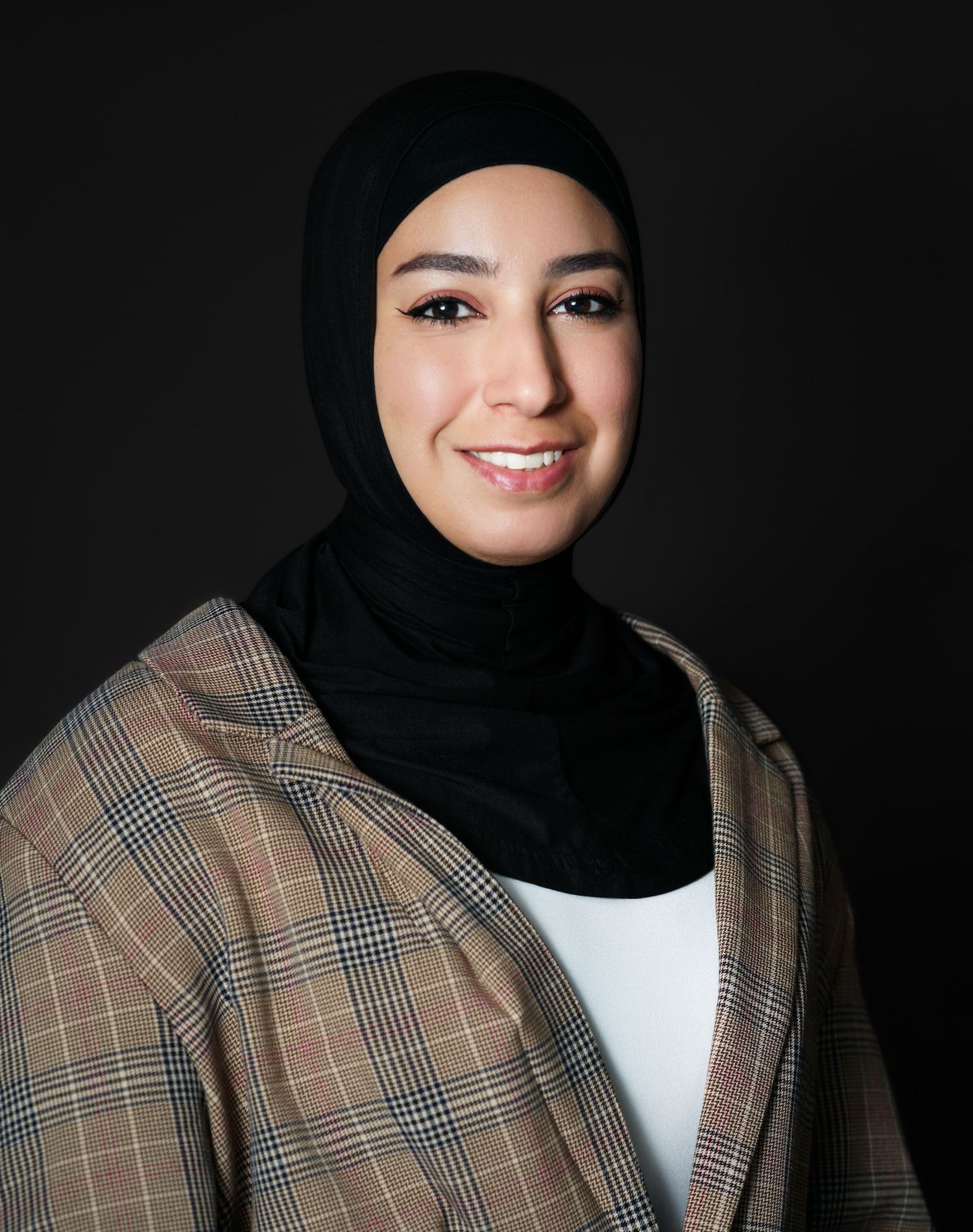 Professional woman wearing black hijab and plaid blazer smiling against dark background.