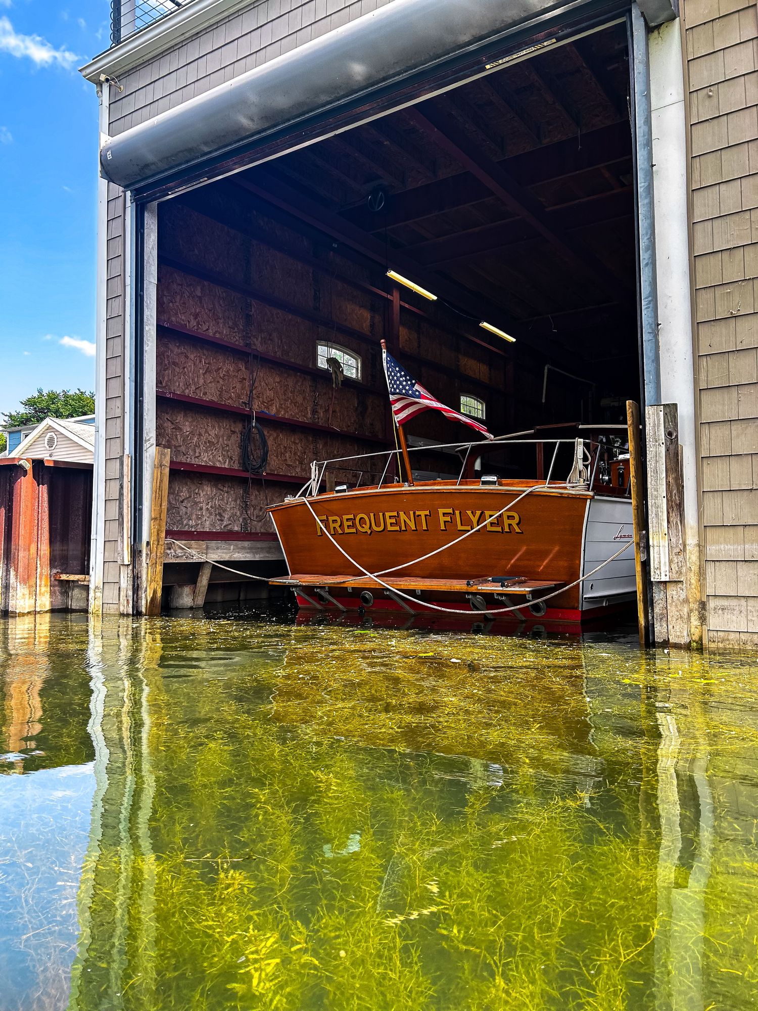 Classic wooden boat named Frequent Flyer docked in a boathouse with an American flag, reflecting on green swamp water.