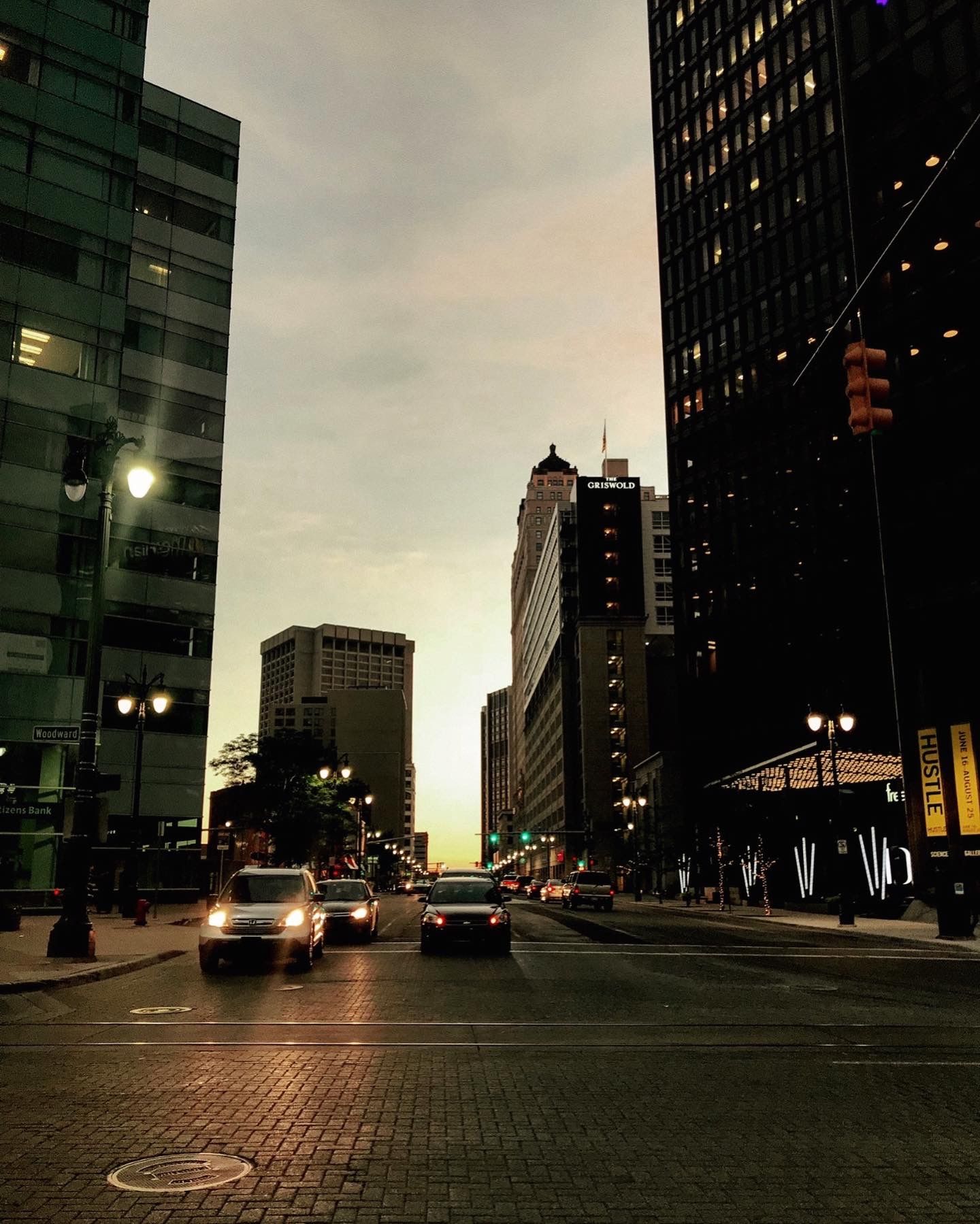 City street at dusk with cars driving between tall buildings, glowing streetlights, and a colorful sunset sky.