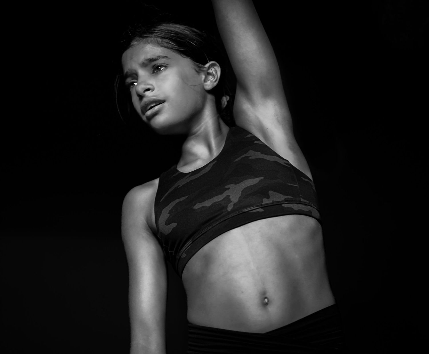 Black and white photo of a young woman in a camo sports bra posing with one arm raised overhead.
