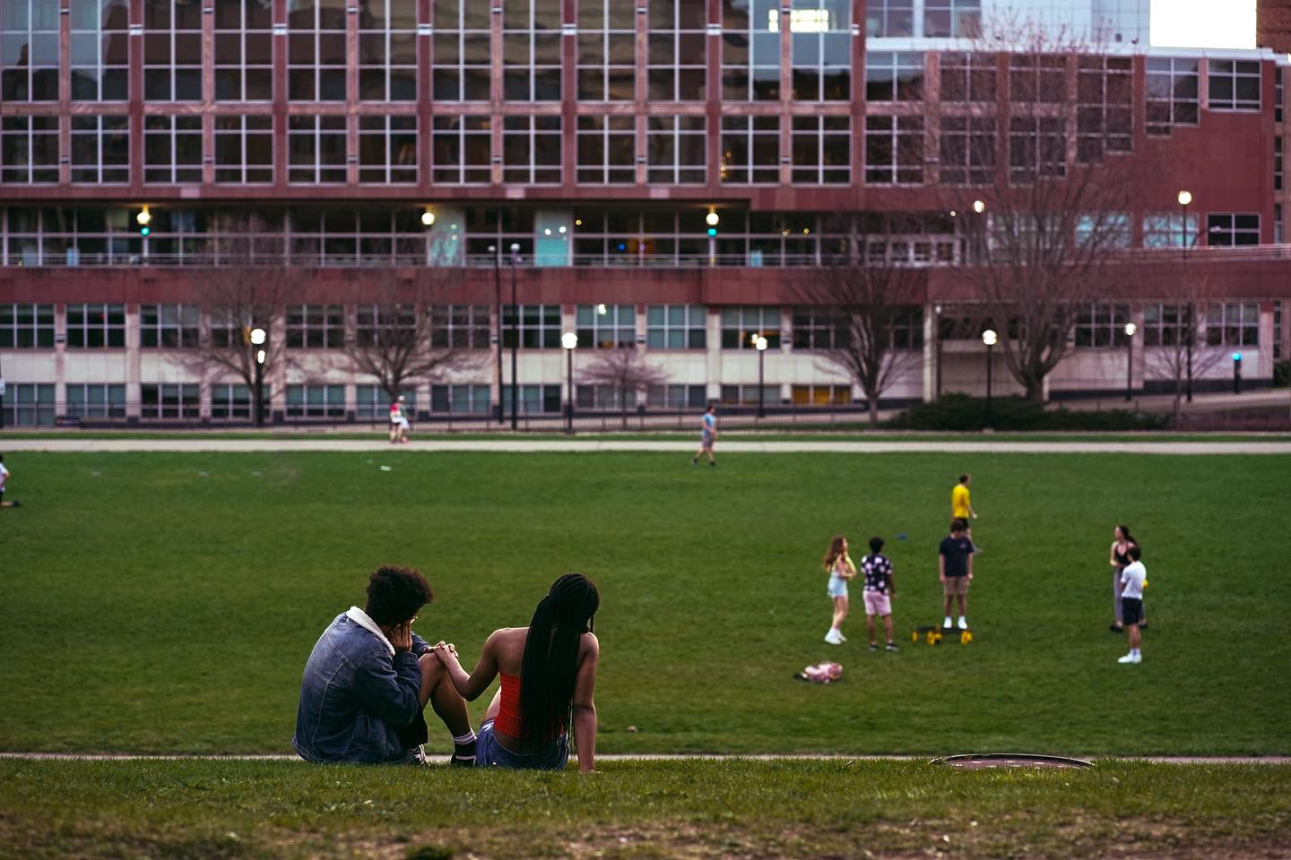 Two people sit on a grassy urban park lawn while children play nearby, with a large brick building in the background.