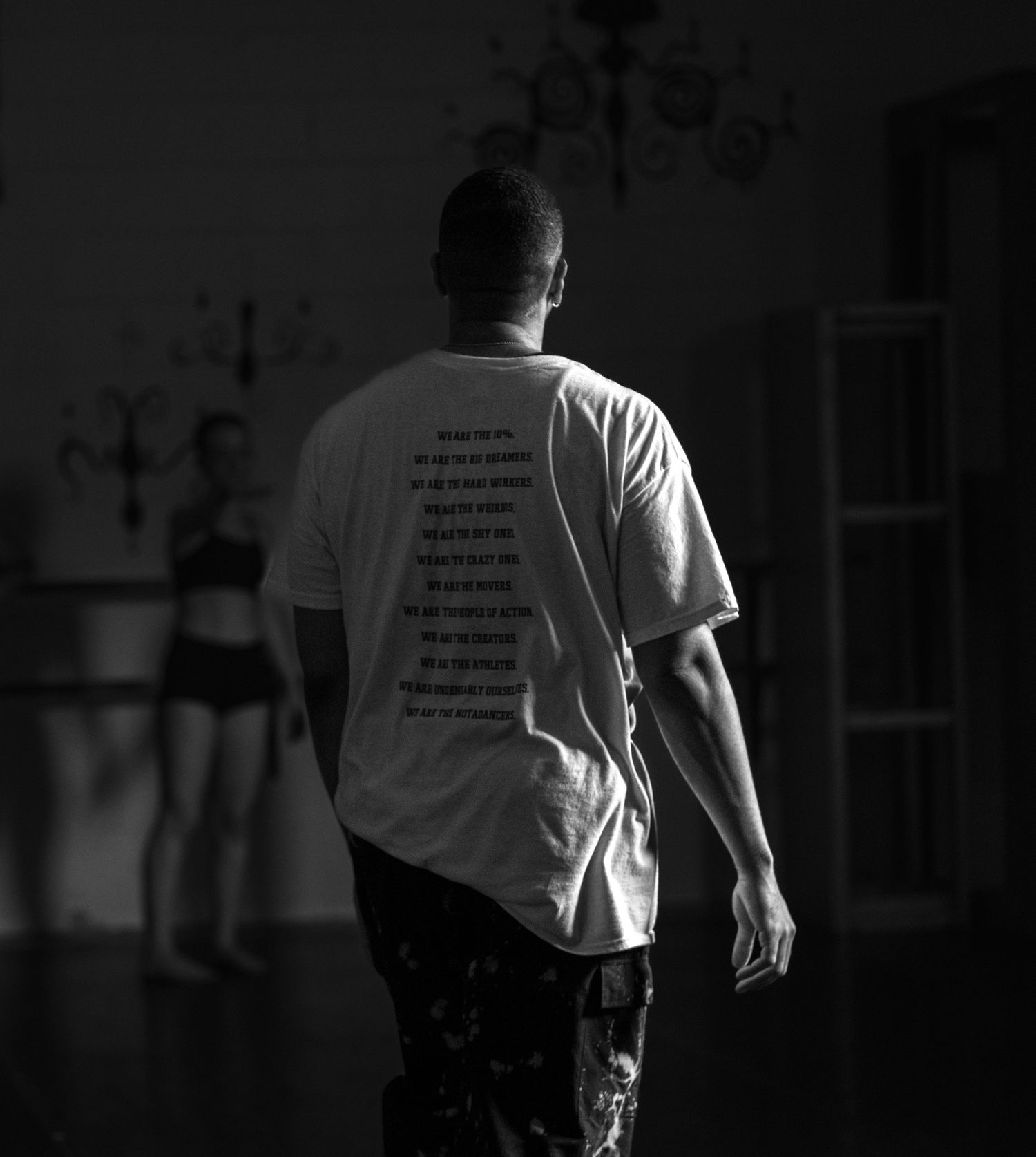 Black and white photo of a man in a graphic t-shirt walking away in a dimly lit dance studio.