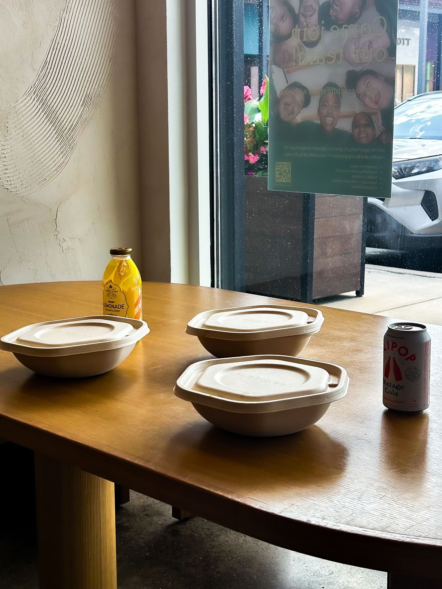 Three covered bowls, a soda can, and juice bottle on a wooden table near a window in a cafe.