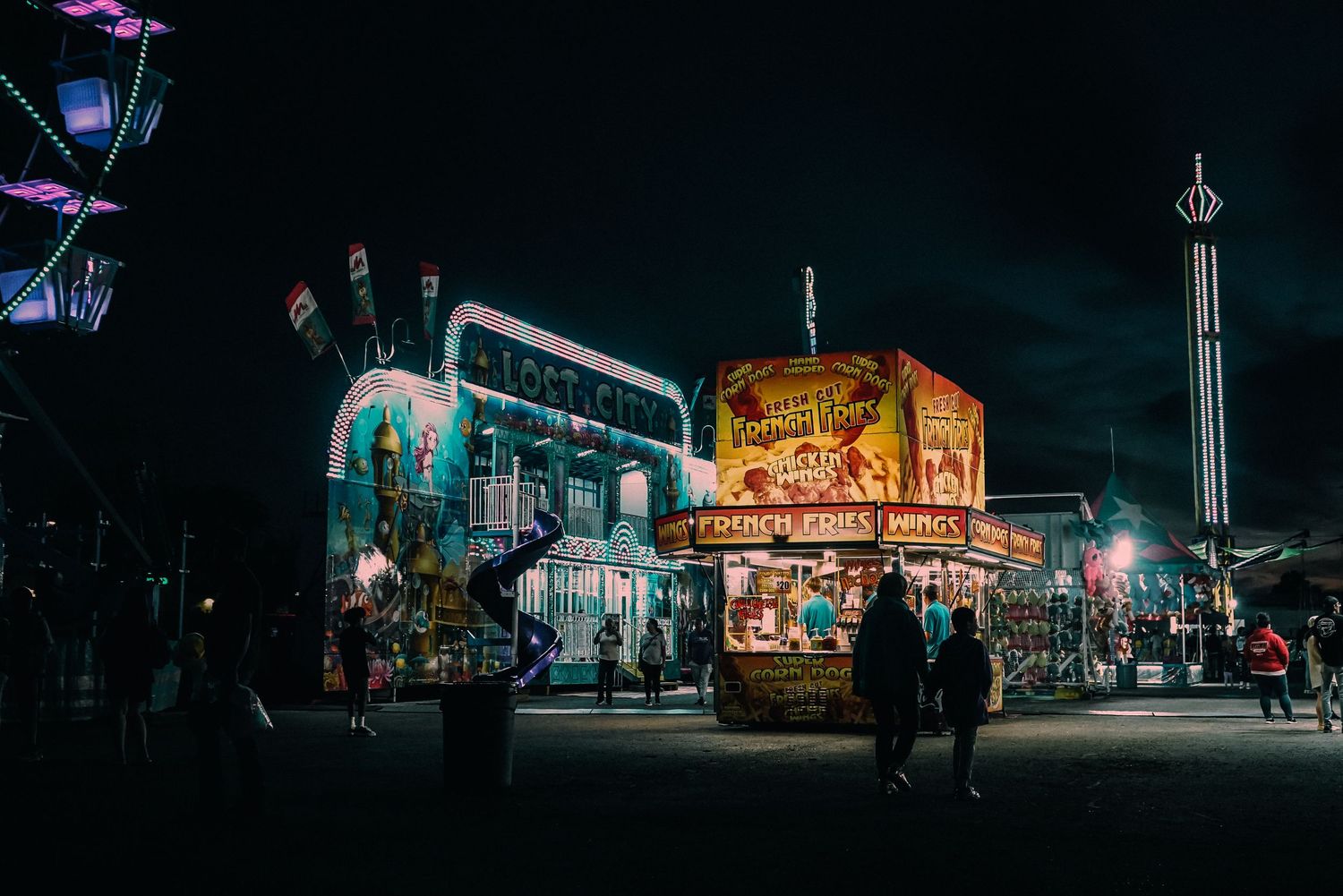 Colorful carnival at night with food stands, Ferris wheel, and thrill rides illuminated against a dark sky.