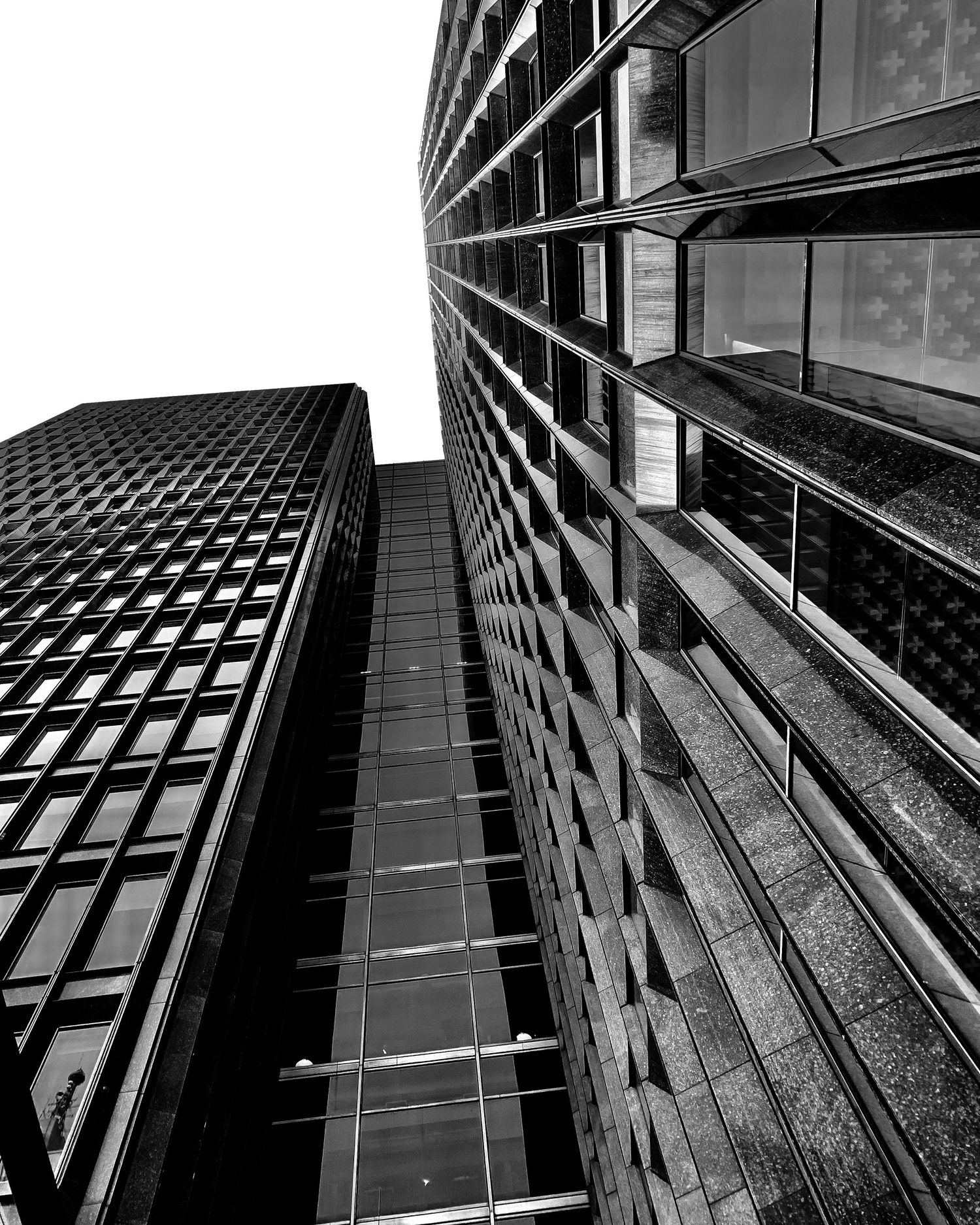 Black and white upward view of towering glass skyscrapers converging against a white sky.