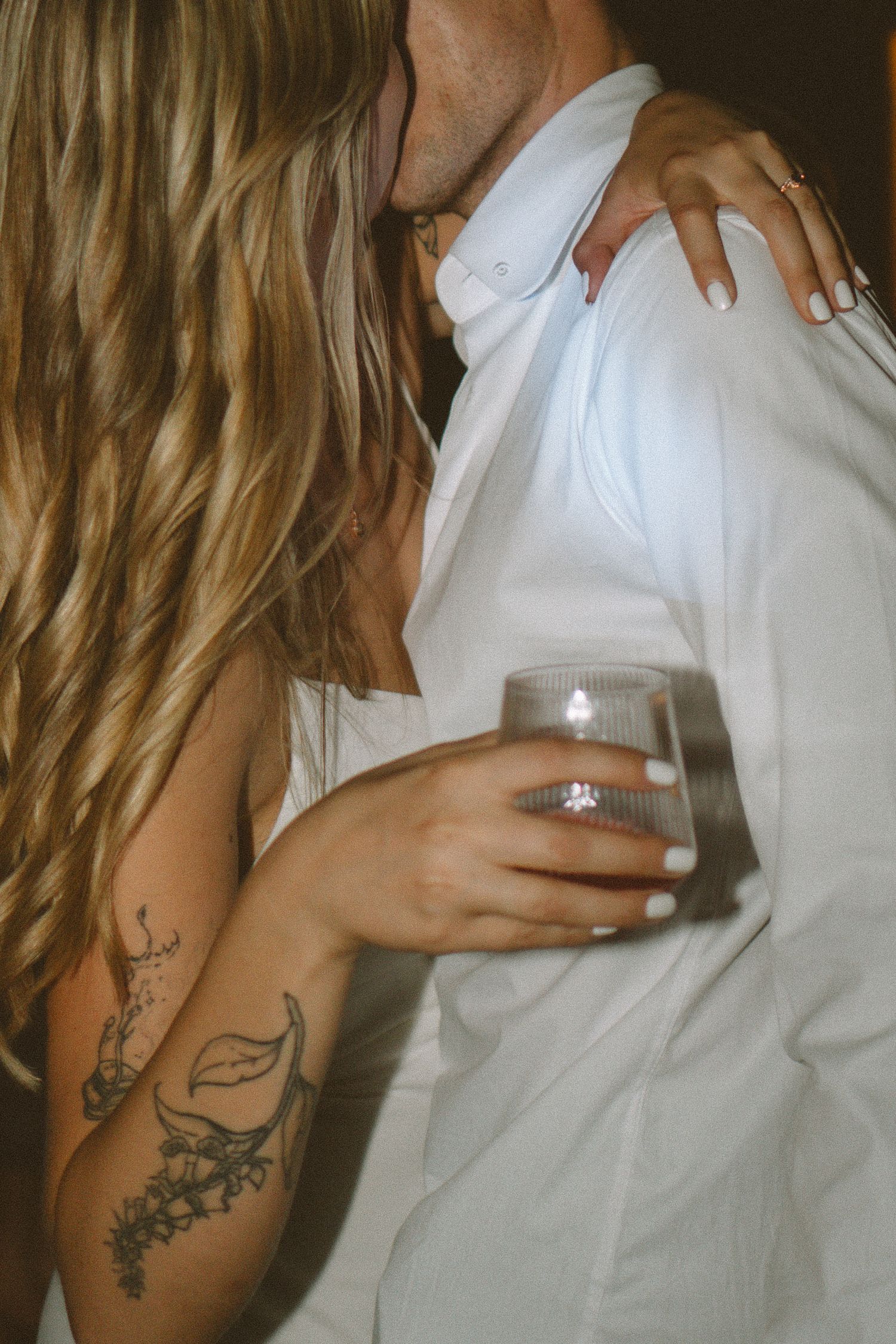 Bride with floral arm tattoo holding whiskey glass while embracing groom in white shirt at reception.