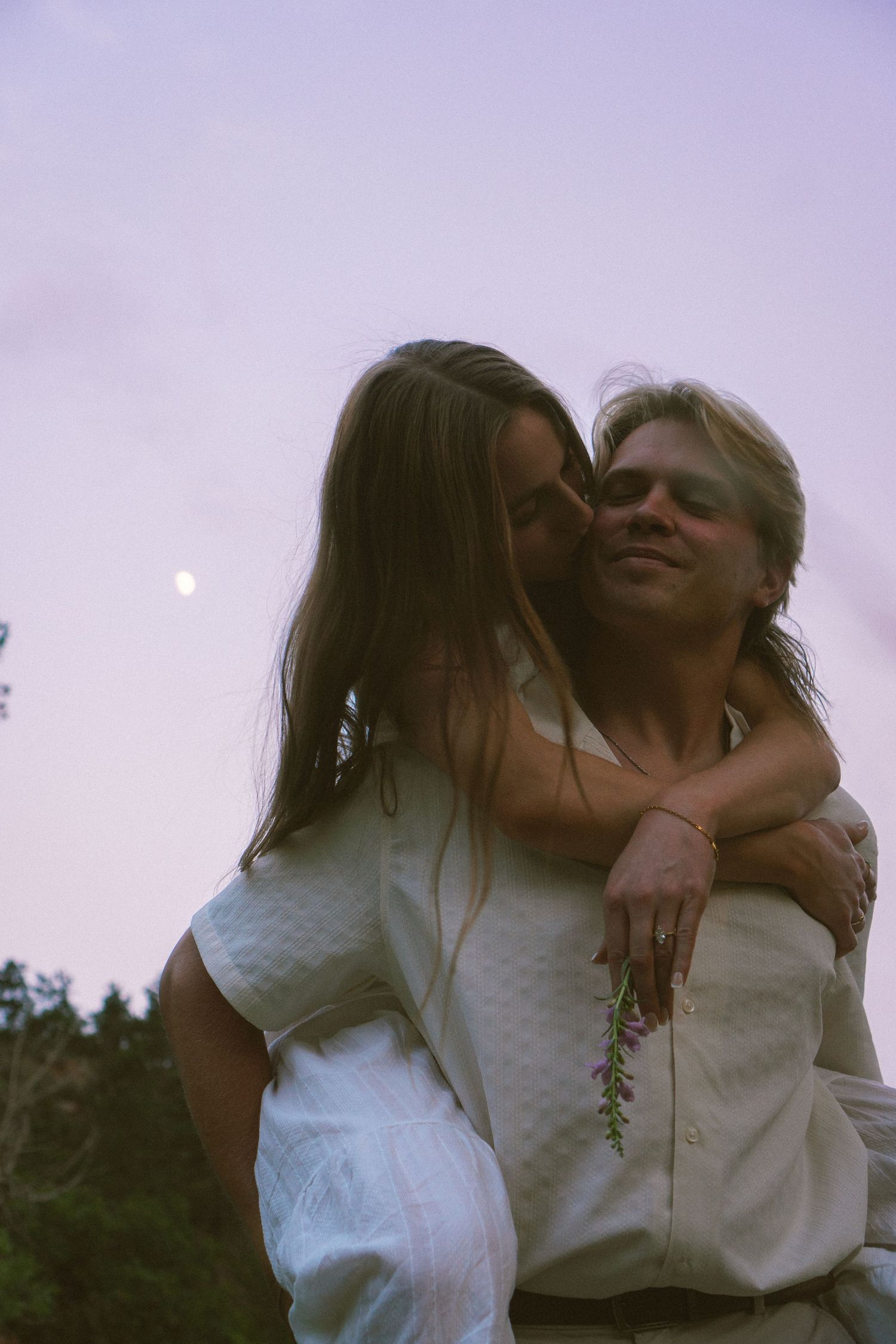 A woman embraces a man from behind kissing his cheek during a soft twilight outdoor session.