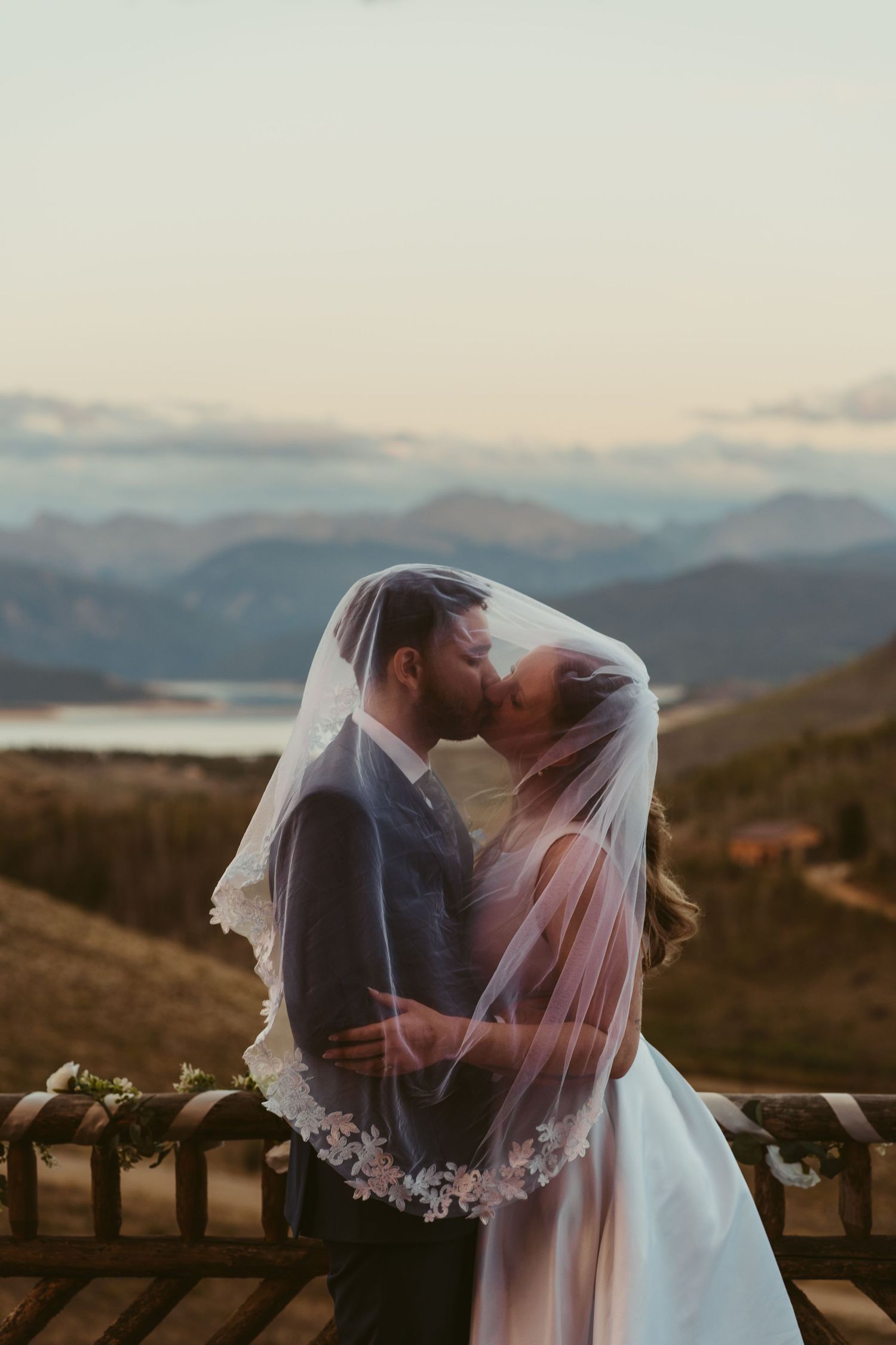 Bride and groom kissing under a lace veil with mountain lake backdrop at sunset wedding.