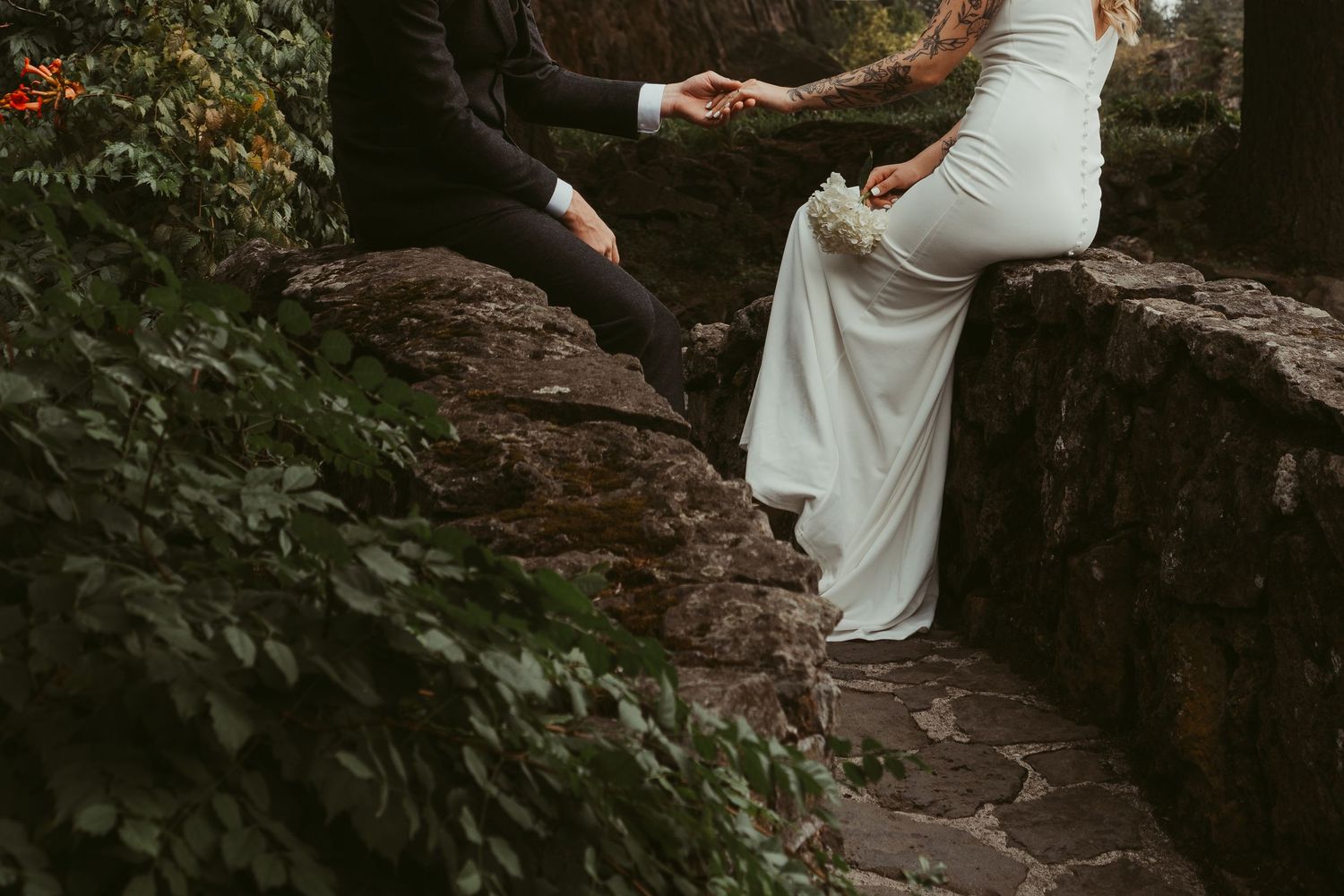 Bride in white gown and groom in black suit hold hands on a stone path surrounded by lush greenery.