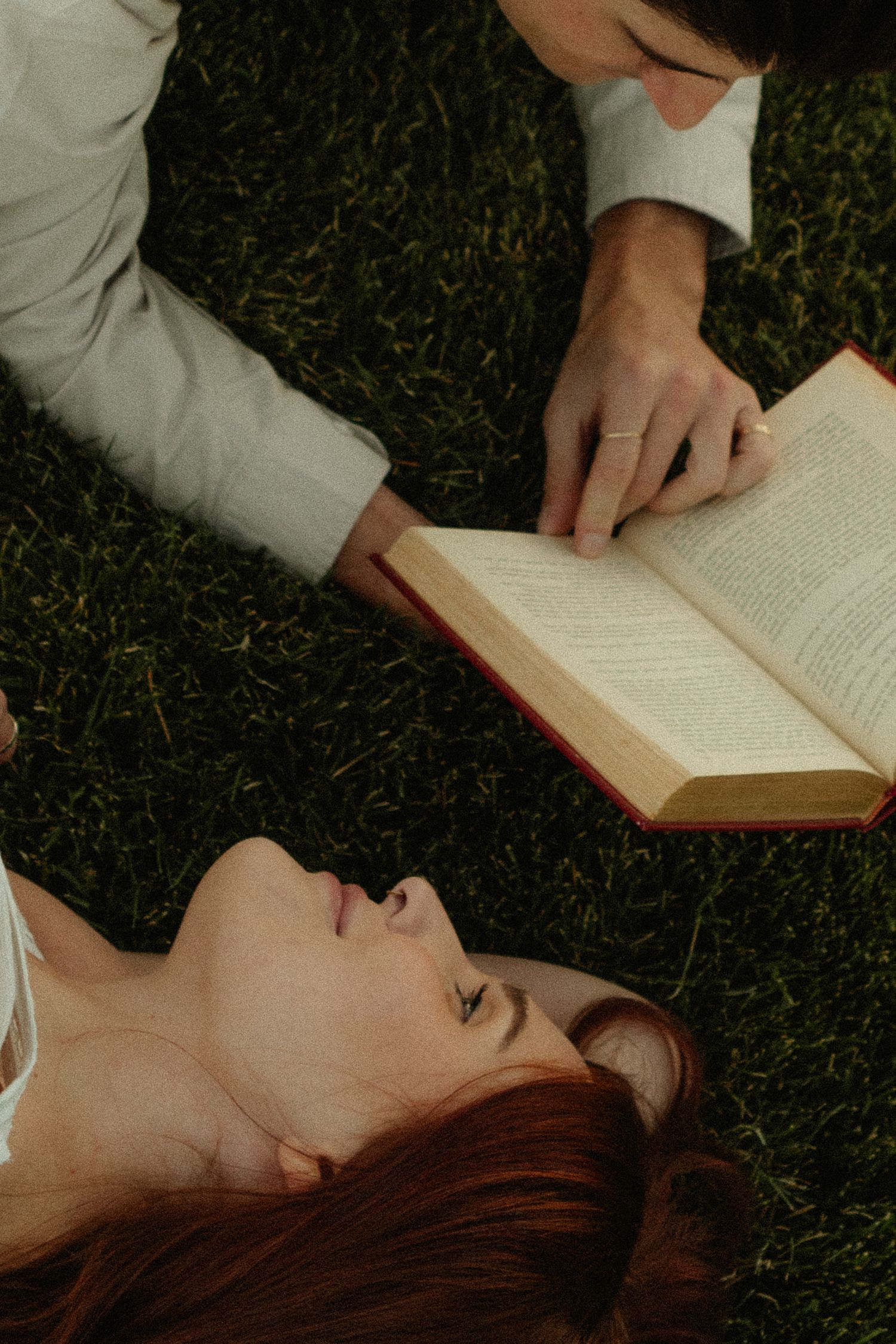 A couple lies on green grass sharing a book, with a redheaded woman looking up and a man pointing at the pages.