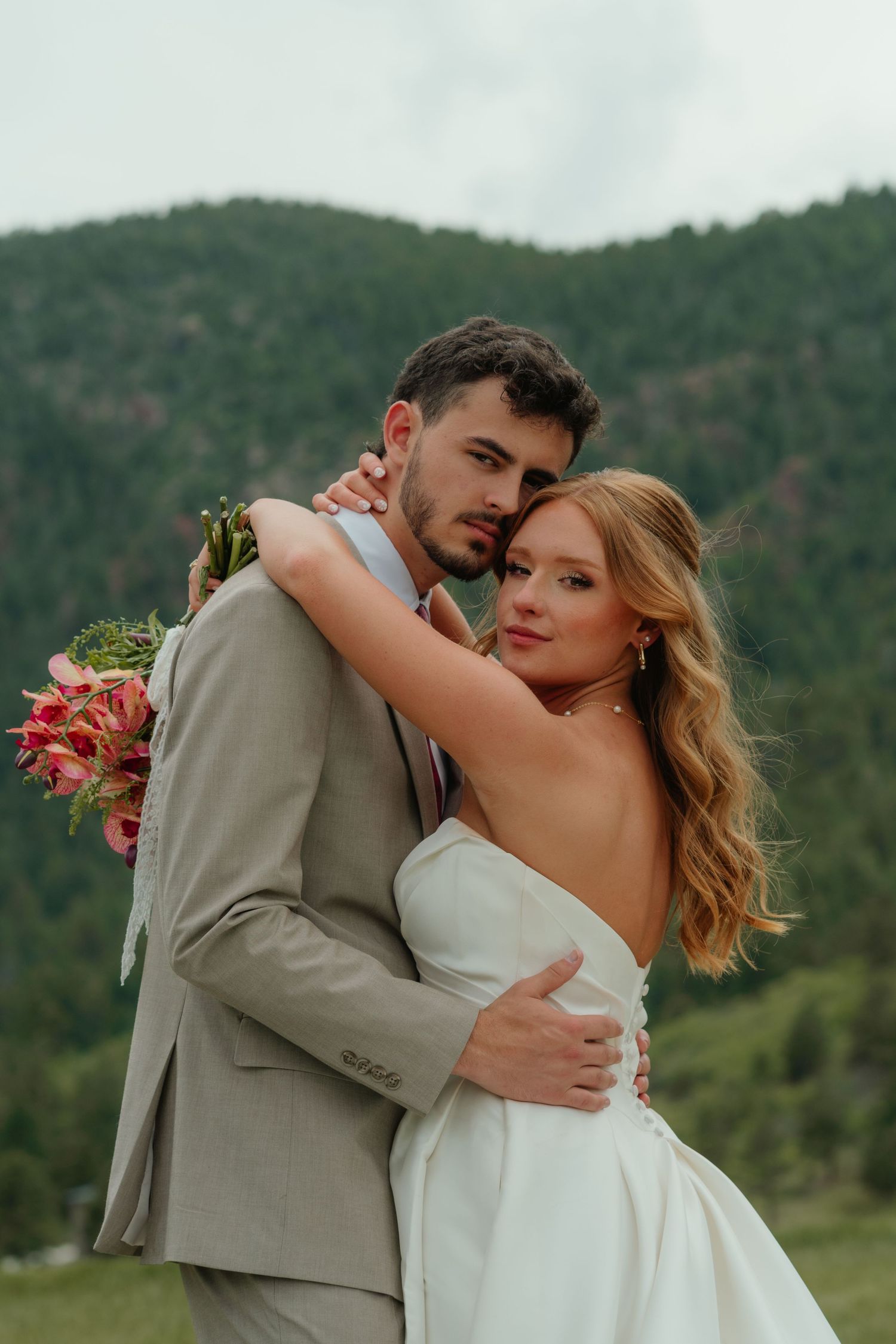 Bride and groom embracing outdoors with mountain backdrop, bride in white gown holding pink bouquet, groom in tan suit.