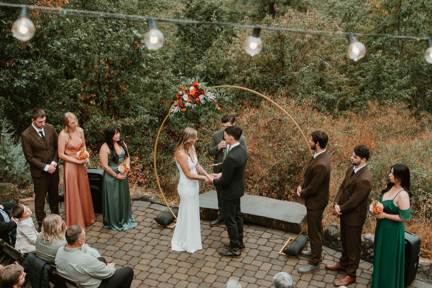 Outdoor wedding ceremony with couple at floral hoop arch, wedding party in green and brown attire on brick patio.