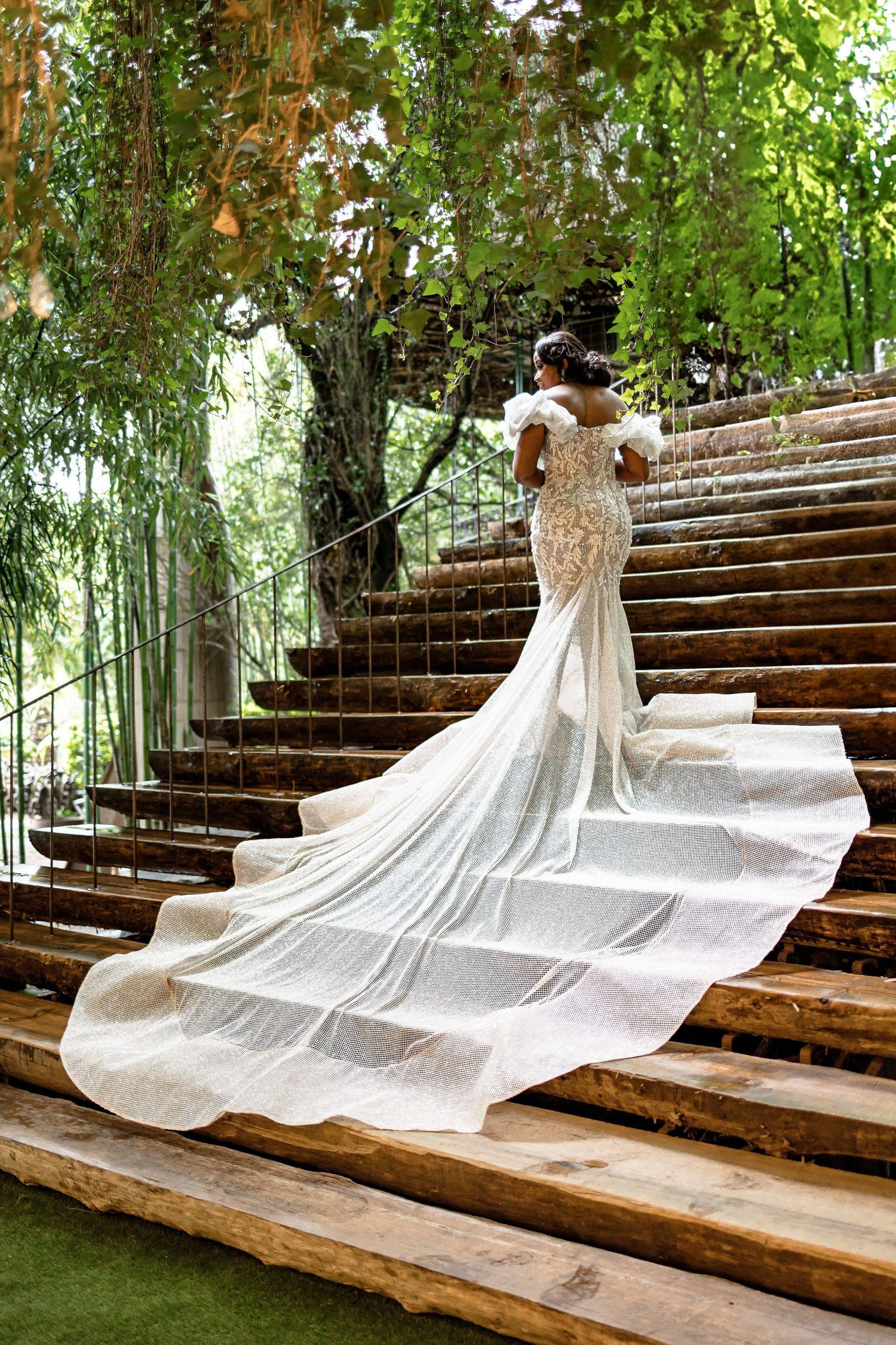 Flowing white wedding gown with long train displayed on rustic wooden steps surrounded by greenery.