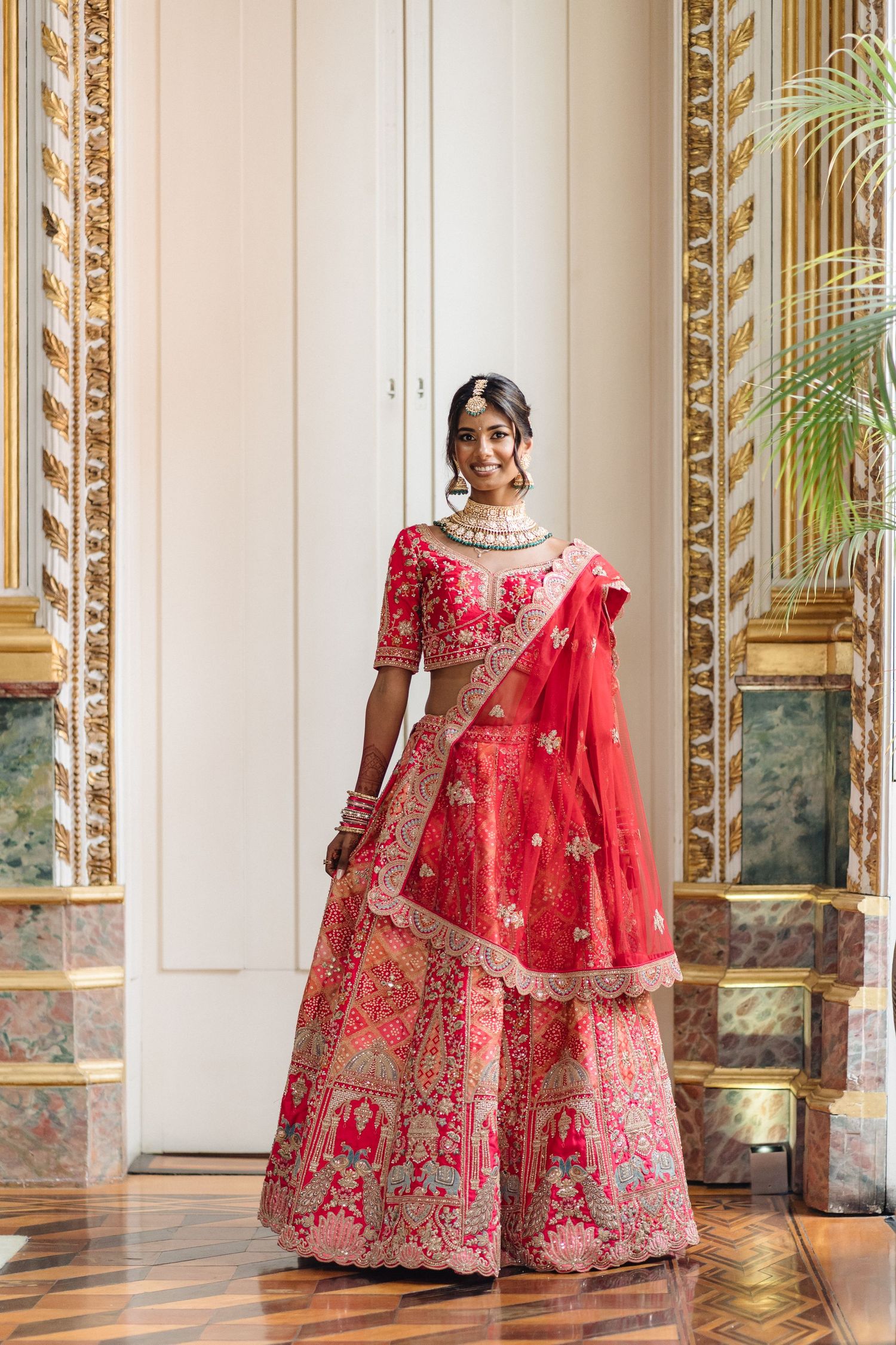 A bride wearing an ornate red and gold embroidered lehenga with traditional Indian bridal jewelry standing in an opulent room.