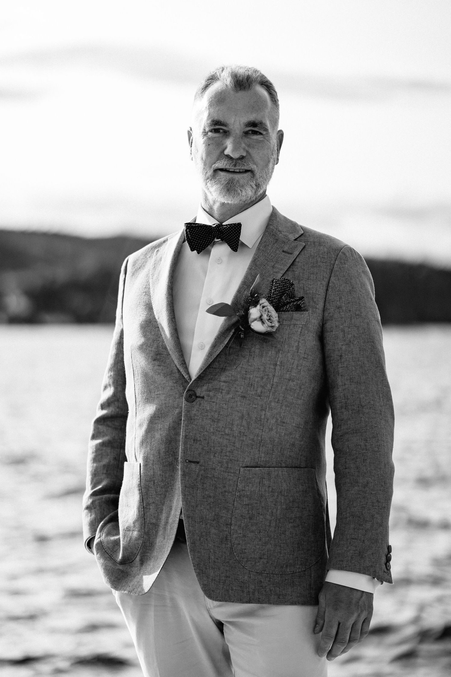 Black and white portrait of groom in grey suit with boutonniere by waterfront.