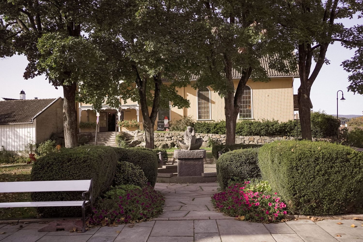 A curved concrete walkway leads through a landscaped garden with trees and bushes on a sunny day.
