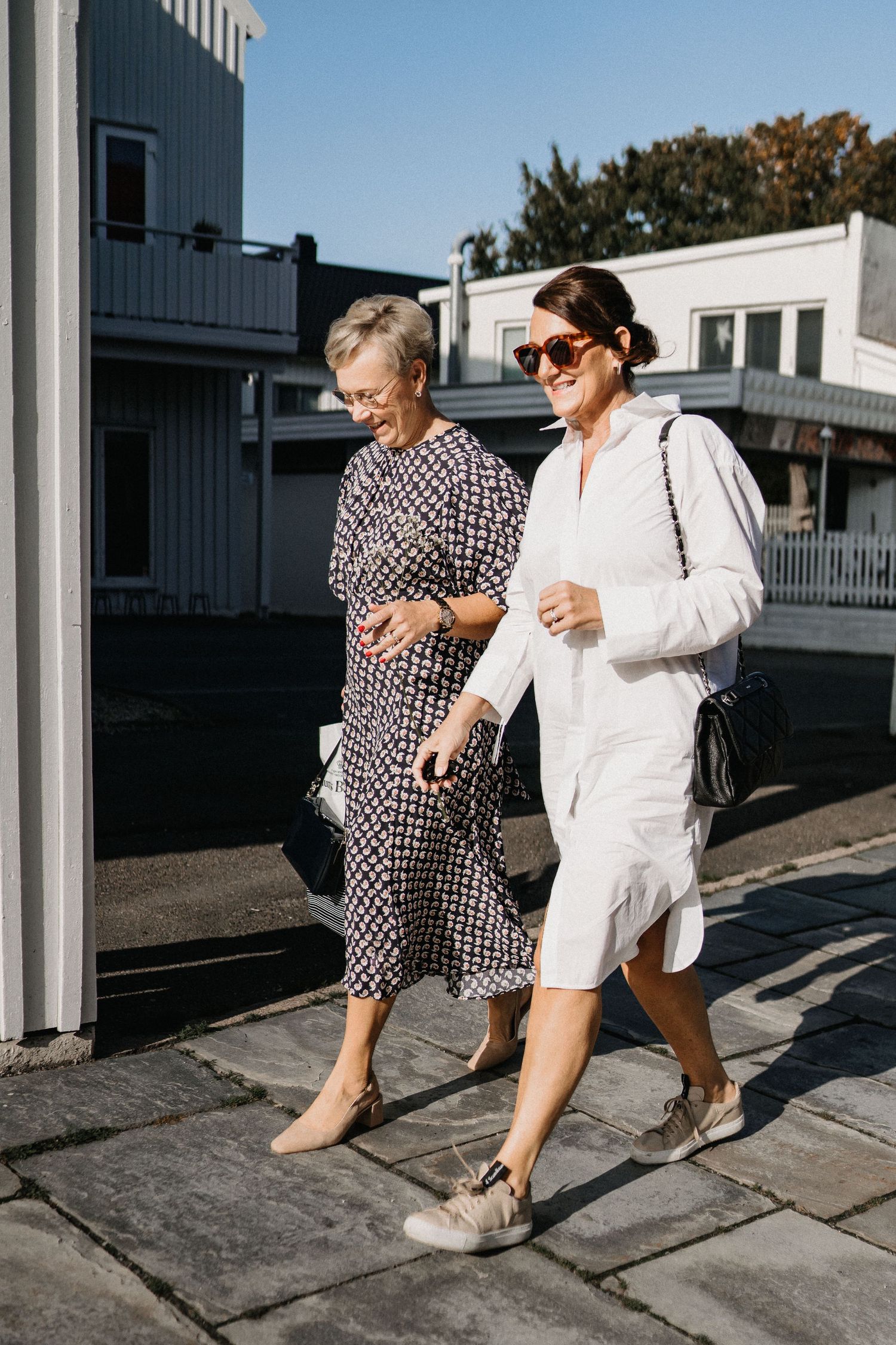 Two people walking together on a street in black and white polka dot and white outfits casting long shadows.