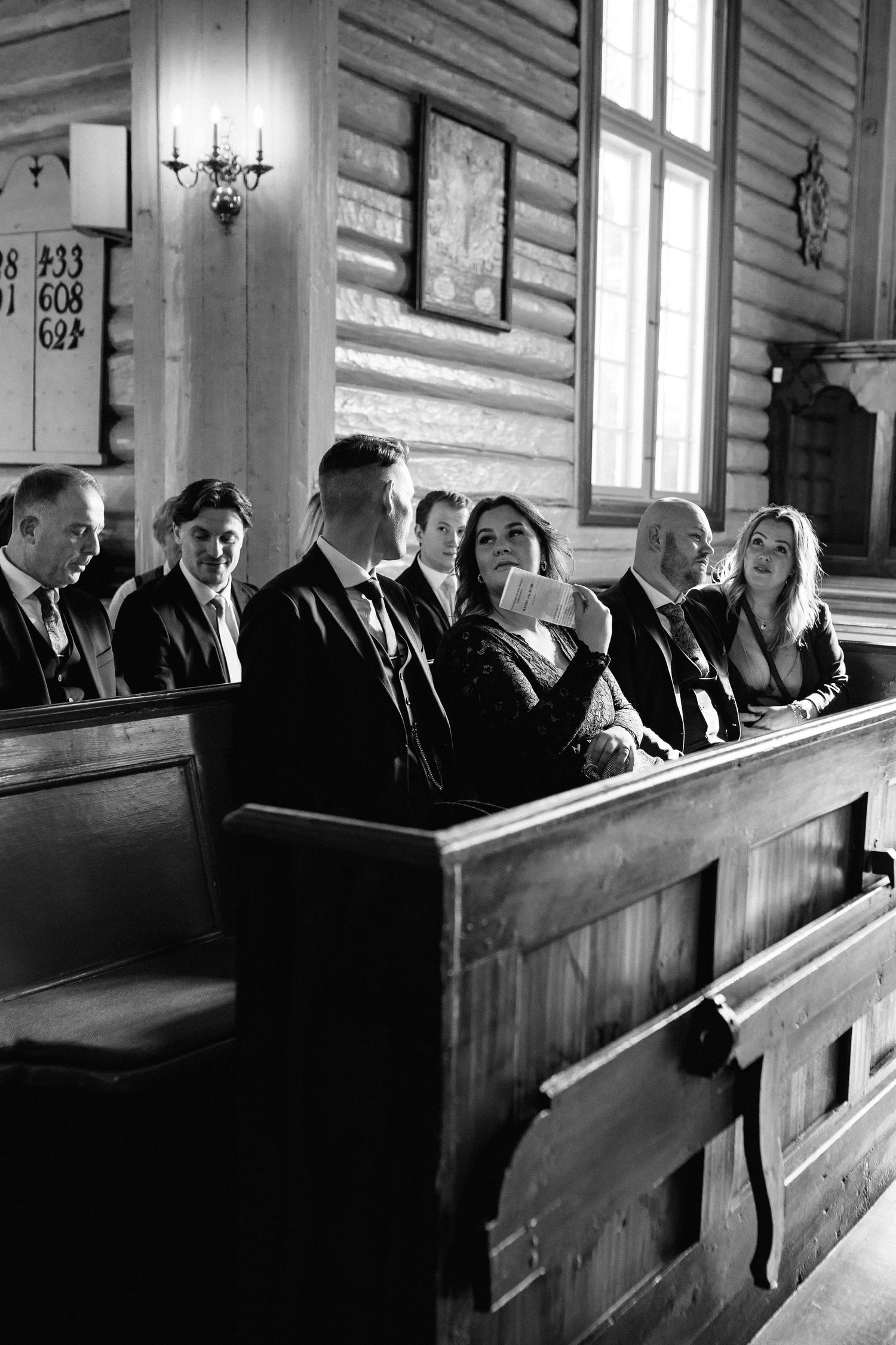Wedding guests seated in traditional wooden church pews captured in dramatic black and white.