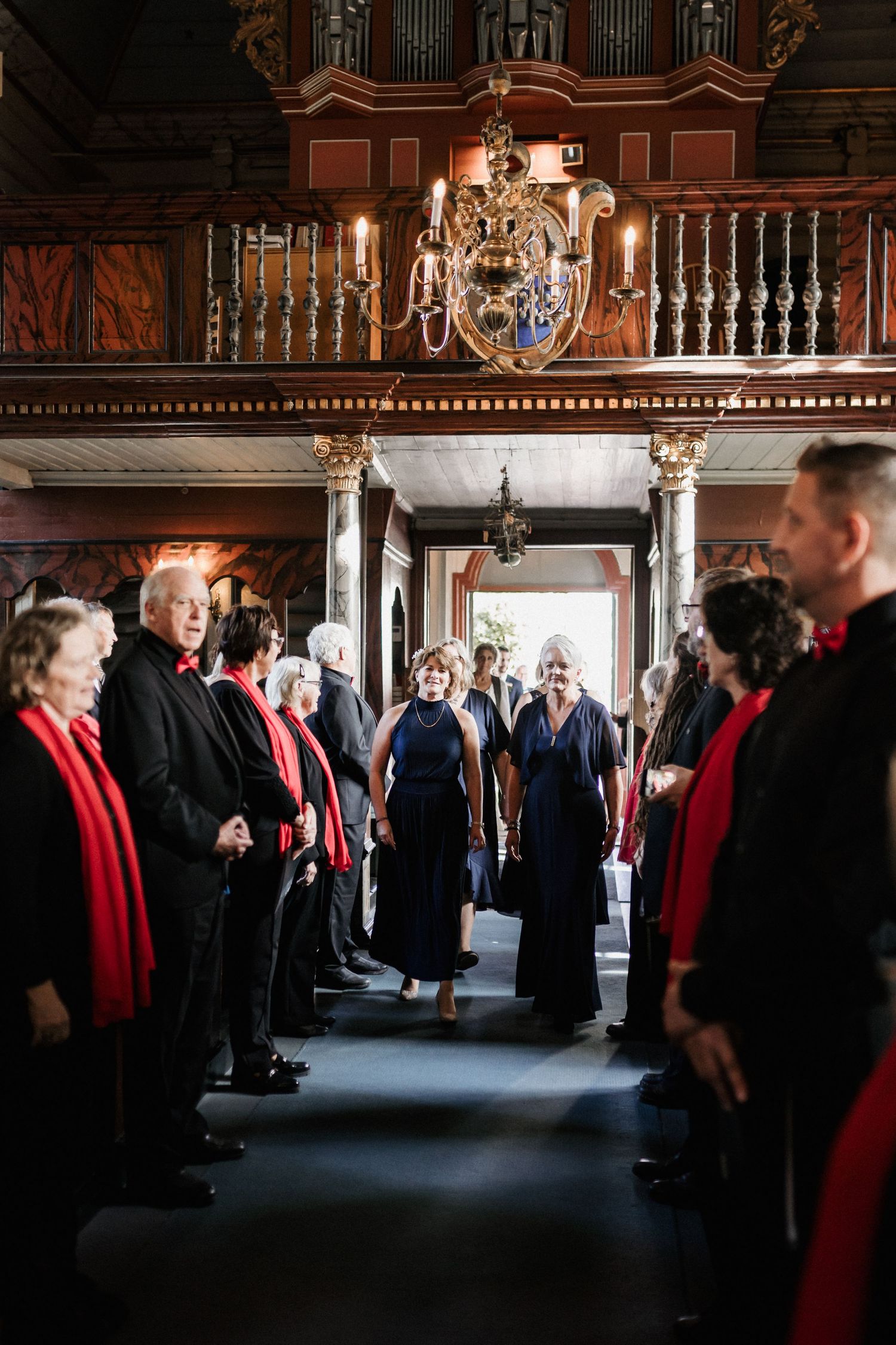 Formal gathering in an ornate hall with people wearing black suits and red robes.