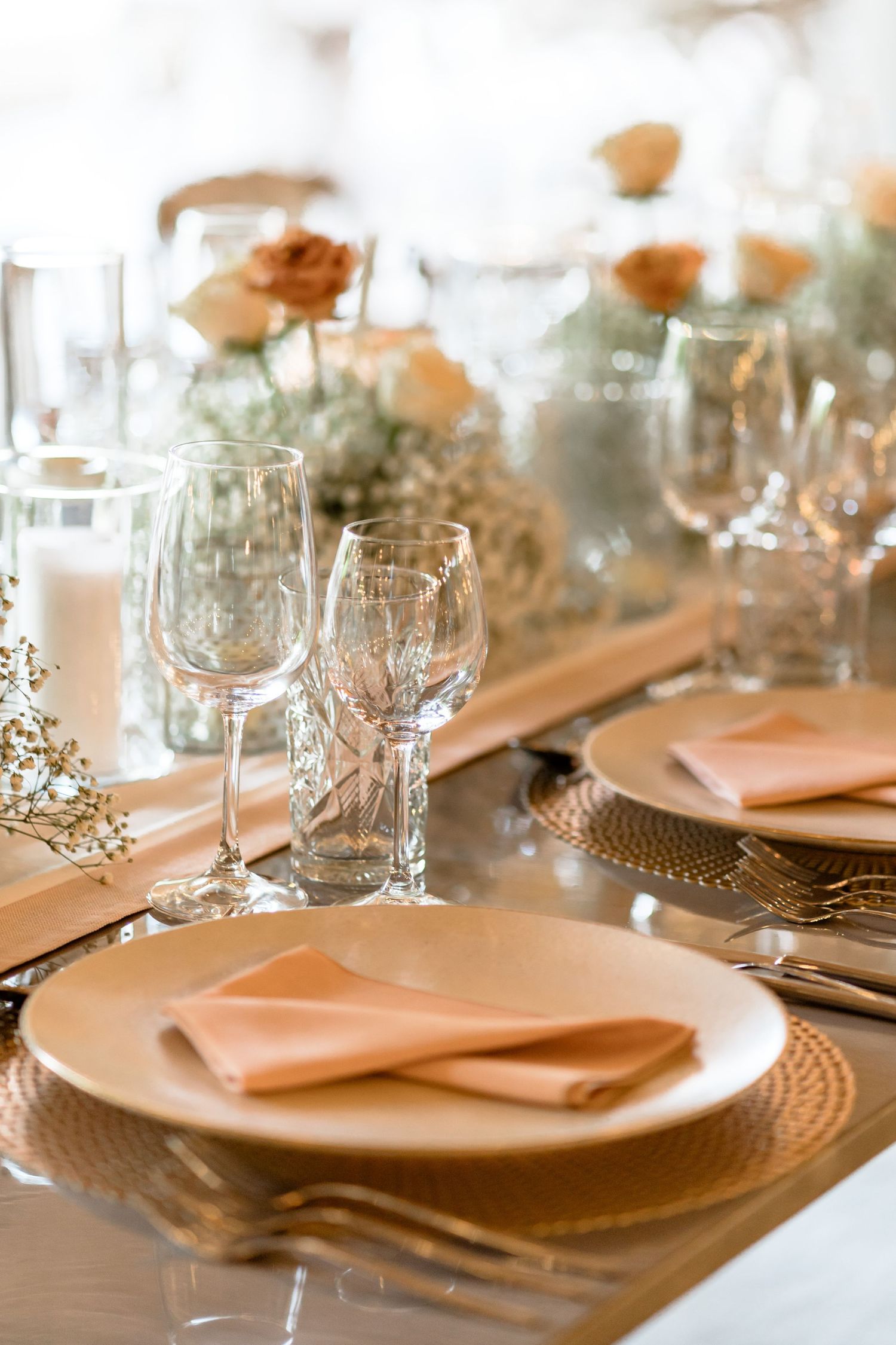 Wedding reception place settings with gold charger plates, crystal glasses, and peach napkins on wooden tables.
