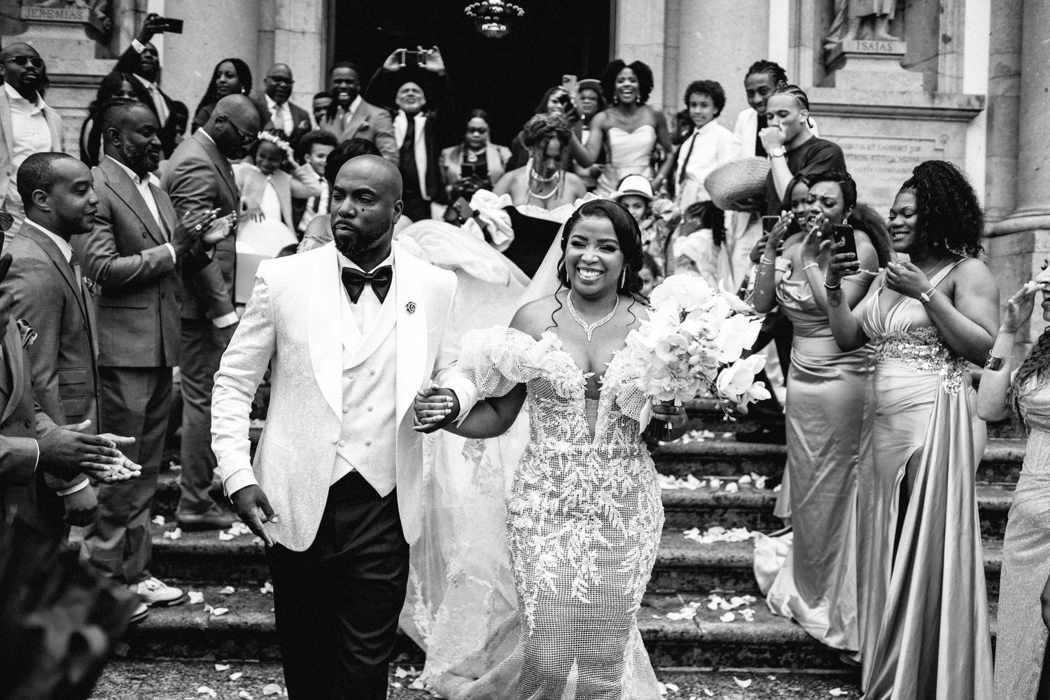 Black and white photo of joyful newlyweds exiting church surrounded by celebrating guests.