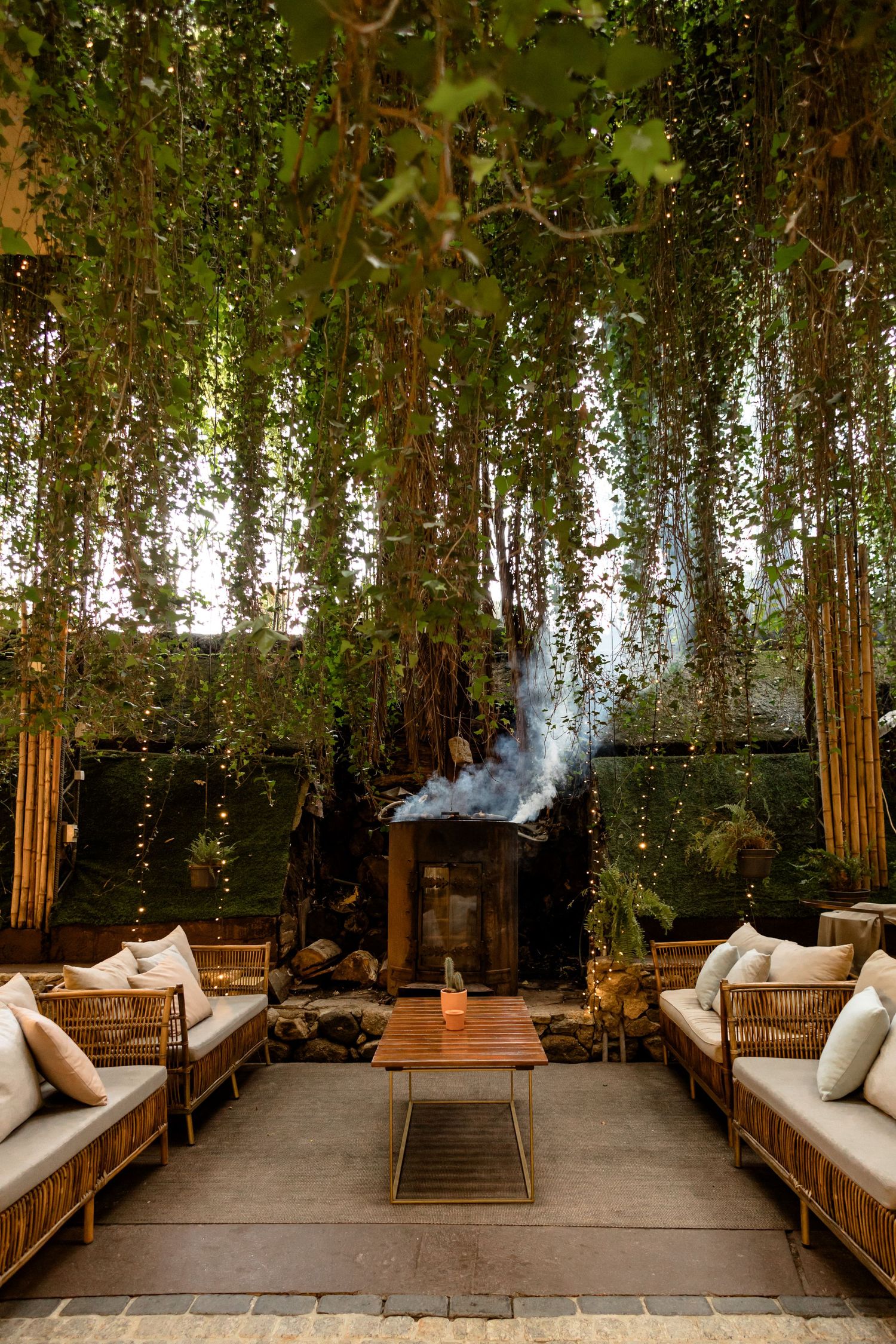 Outdoor lounge area with hanging greenery, wooden benches, and fireplace creates cozy atmosphere.