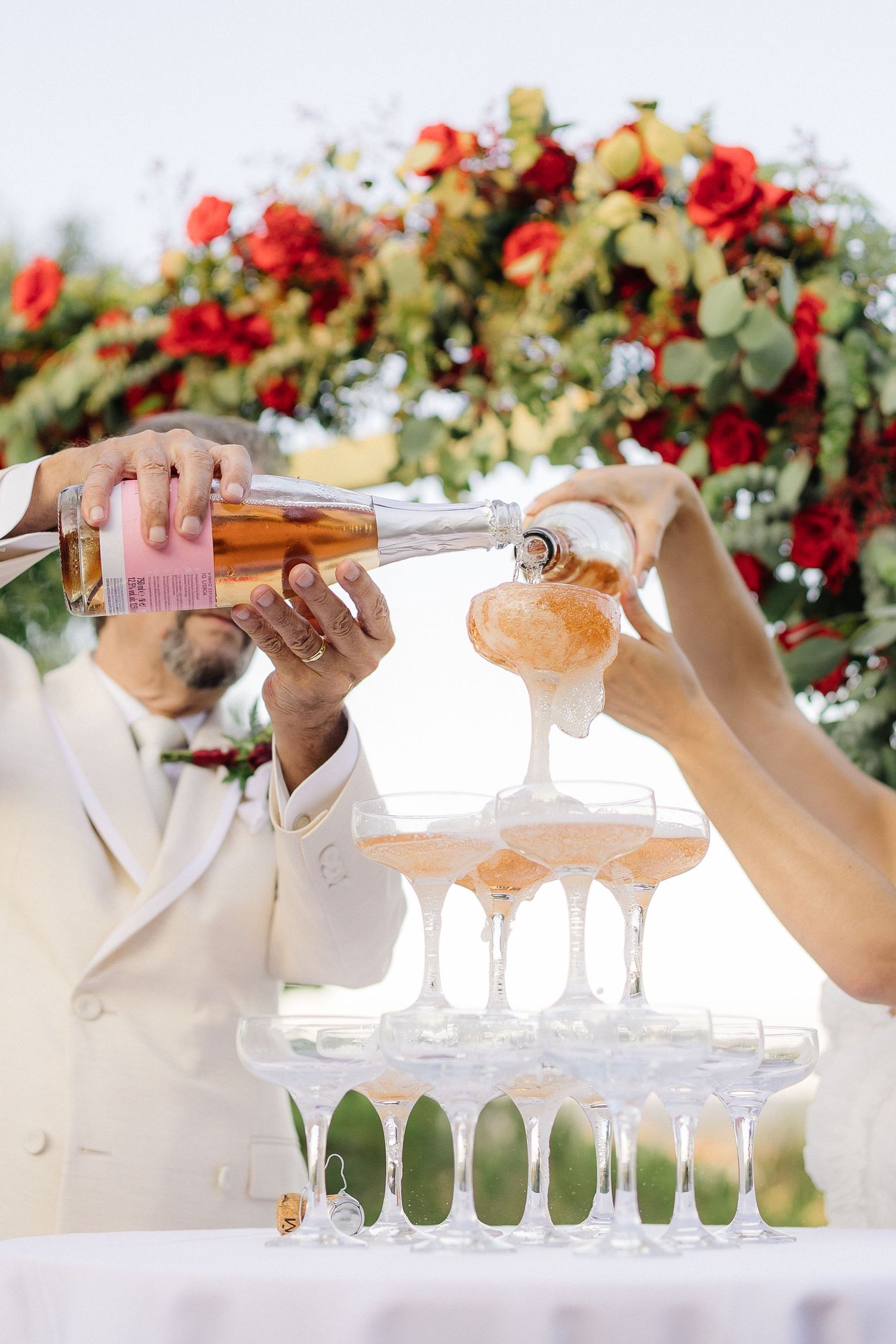 Hands pouring champagne into a tower of crystal glasses with red floral decorations.