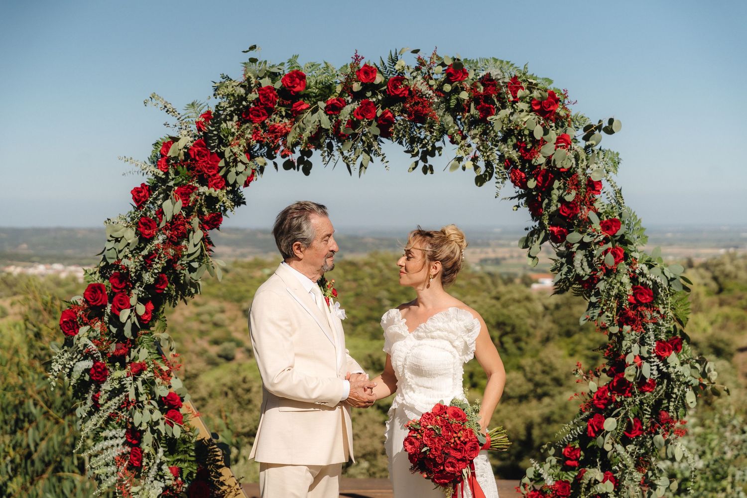 Couple sharing intimate moment under red rose-covered arch with scenic valley view.