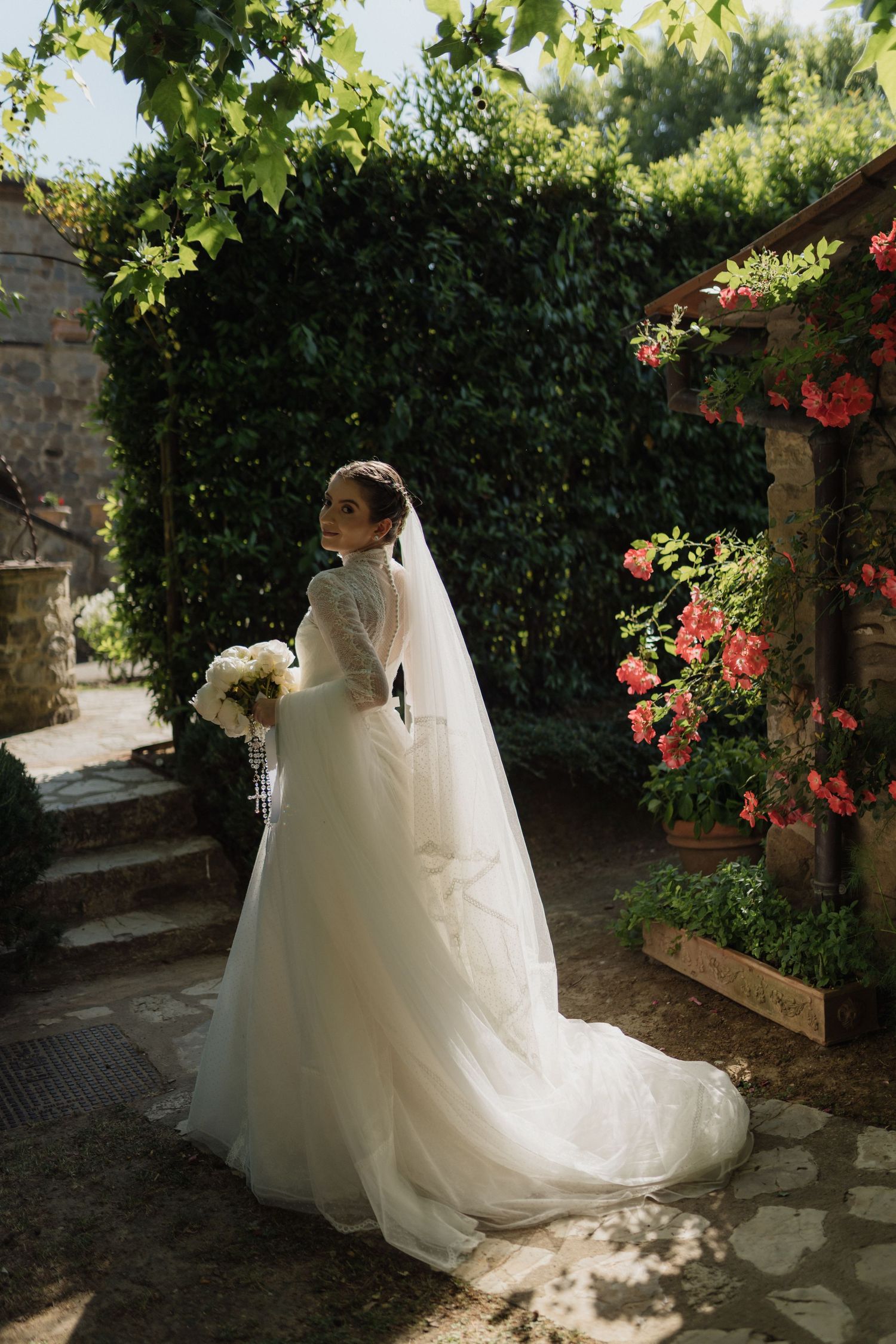 Bride in elegant white tulle gown with veil holding bouquet in lush garden with blooming flowers.
