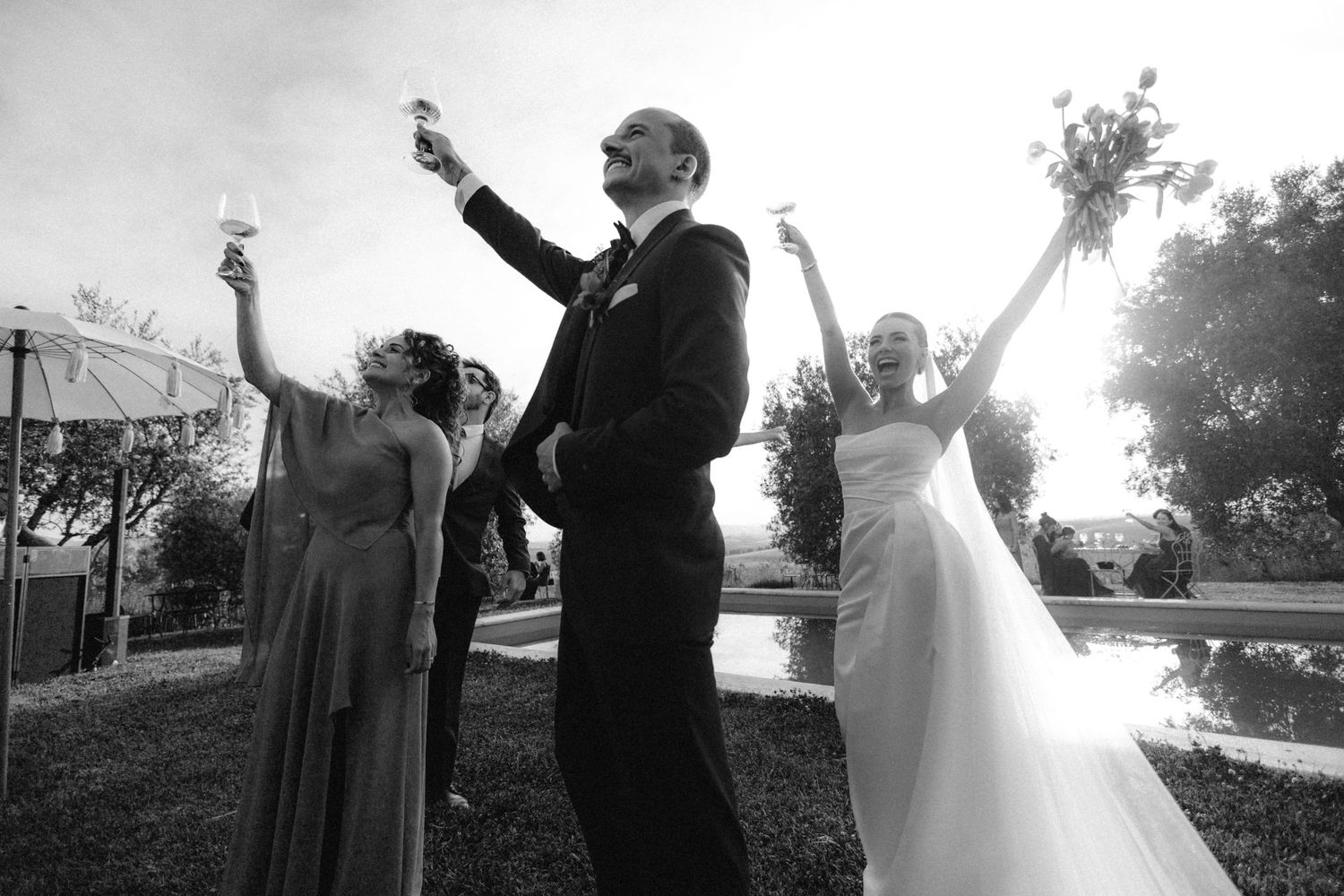 Black and white photo of bride and groom celebrating with raised glasses outdoors at their wedding.