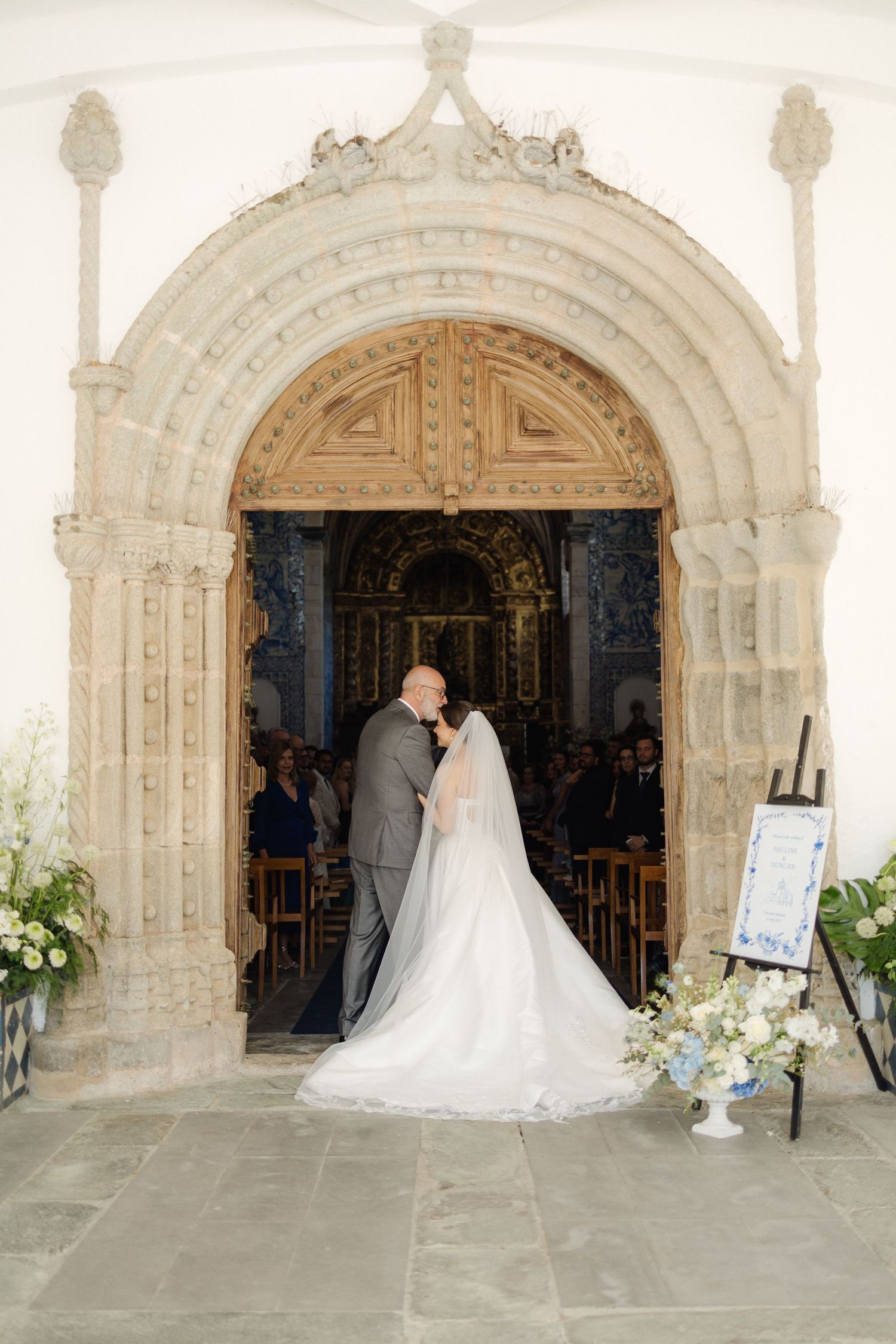 Wedding couple stands in front of a grand stone church archway decorated with white flowers.