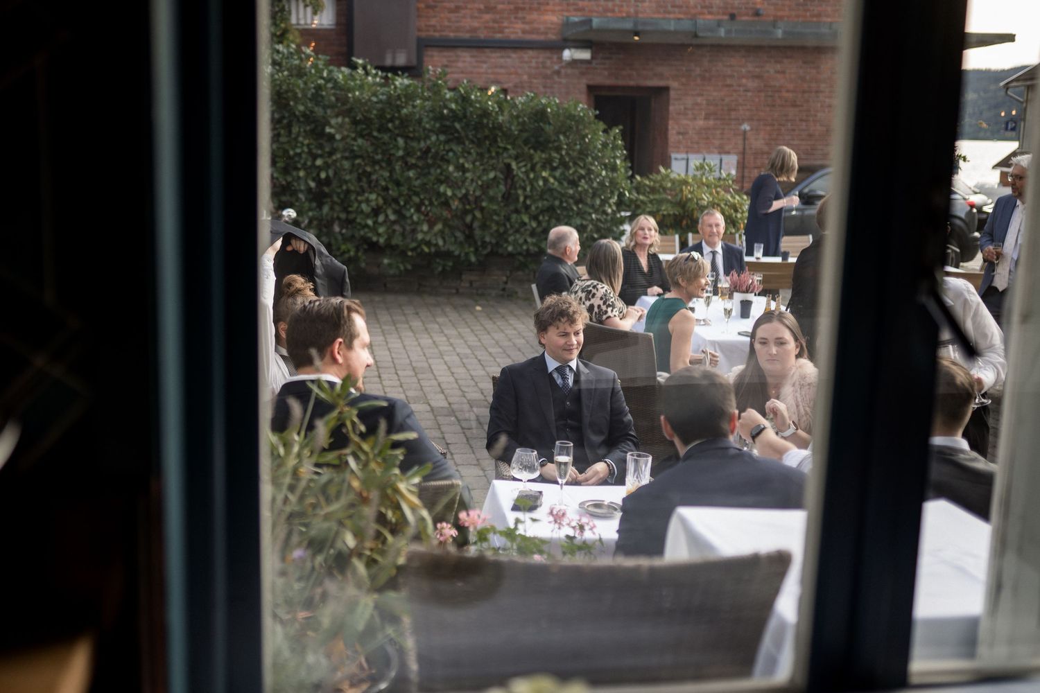 Wedding guests mingle at outdoor reception tables viewed through a window frame.
