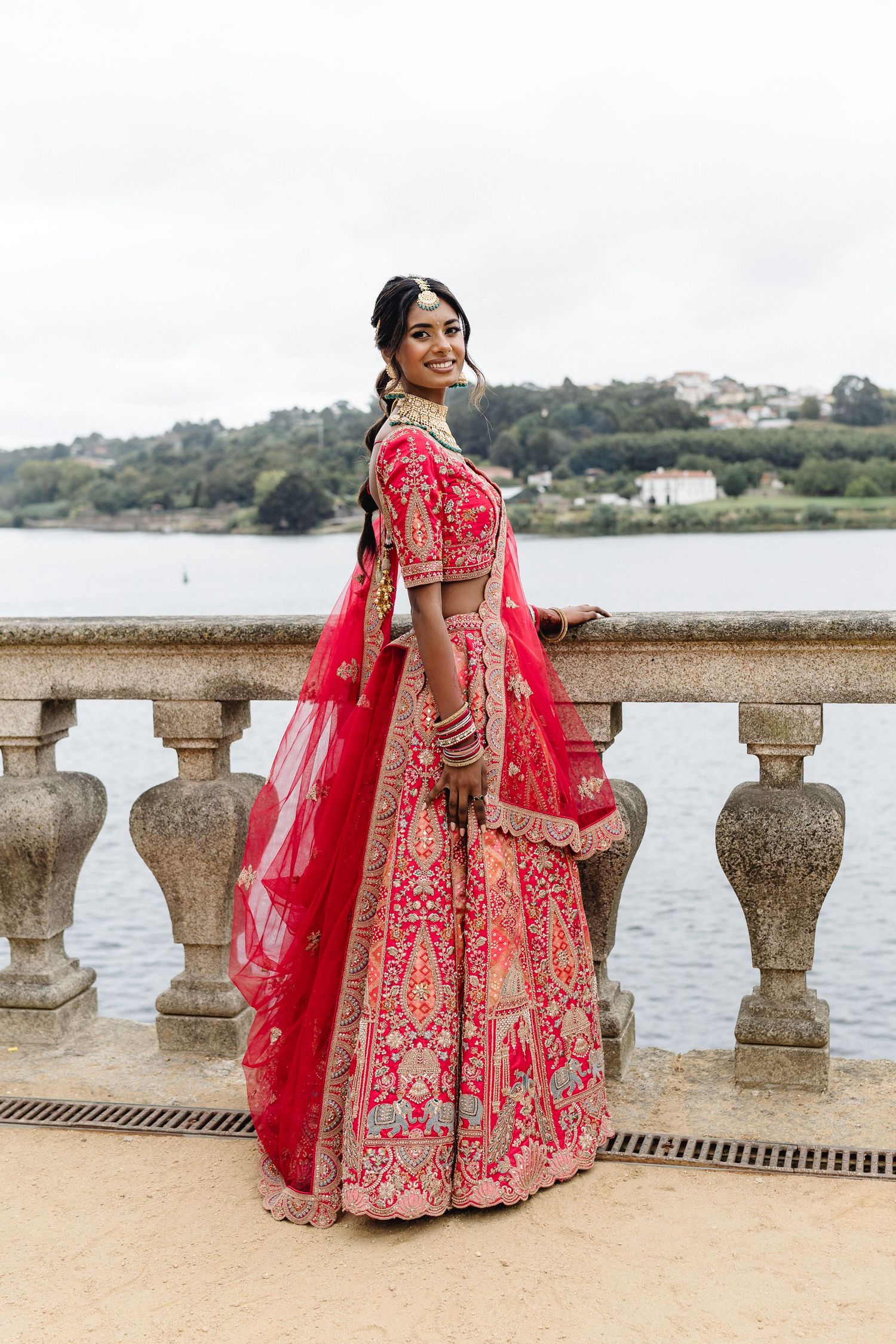 A stunning red and gold embroidered lehenga photographed against a waterfront stone balustrade.