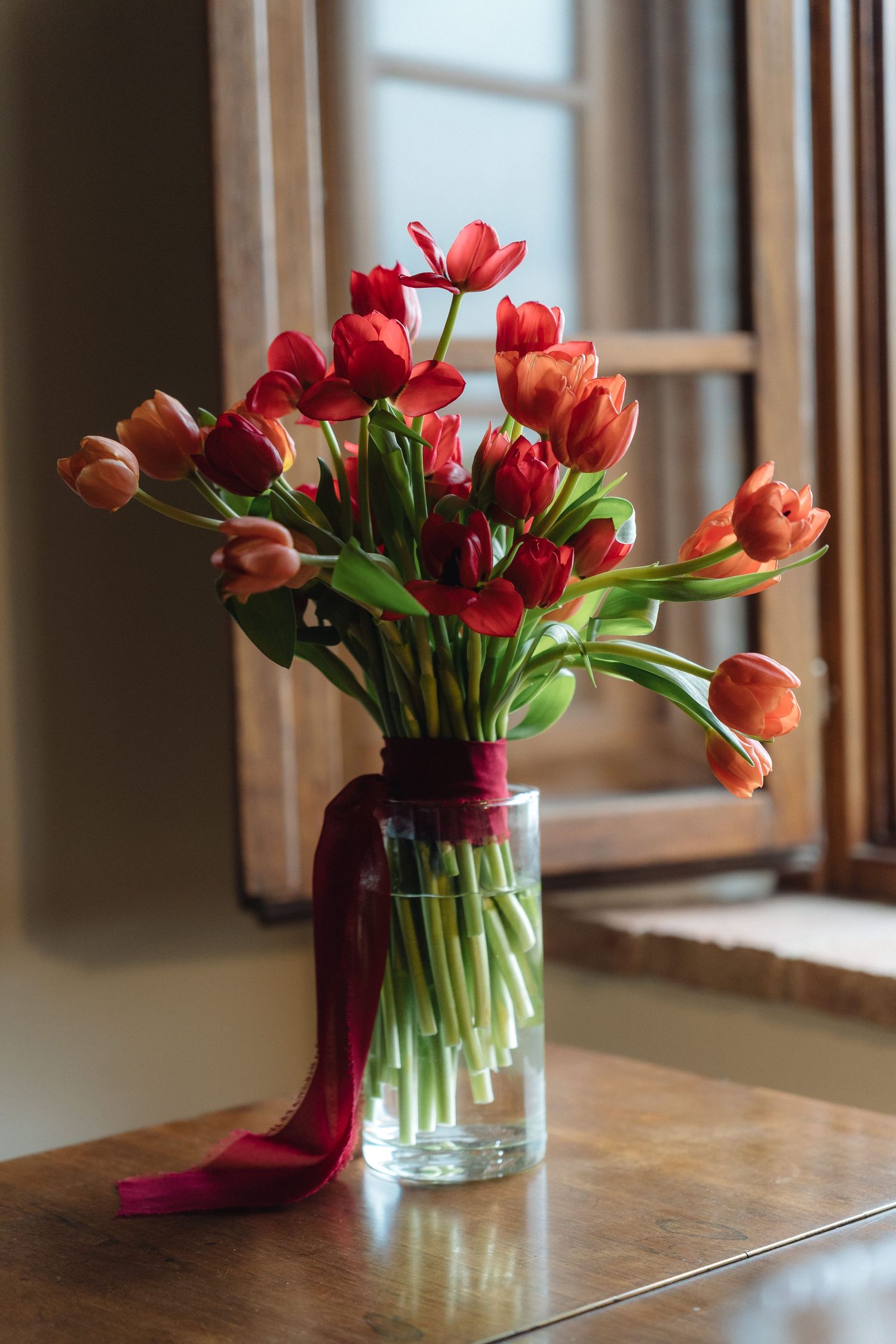 Red and orange tulips in a glass vase tied with burgundy ribbon on a wooden table by a sunlit window.