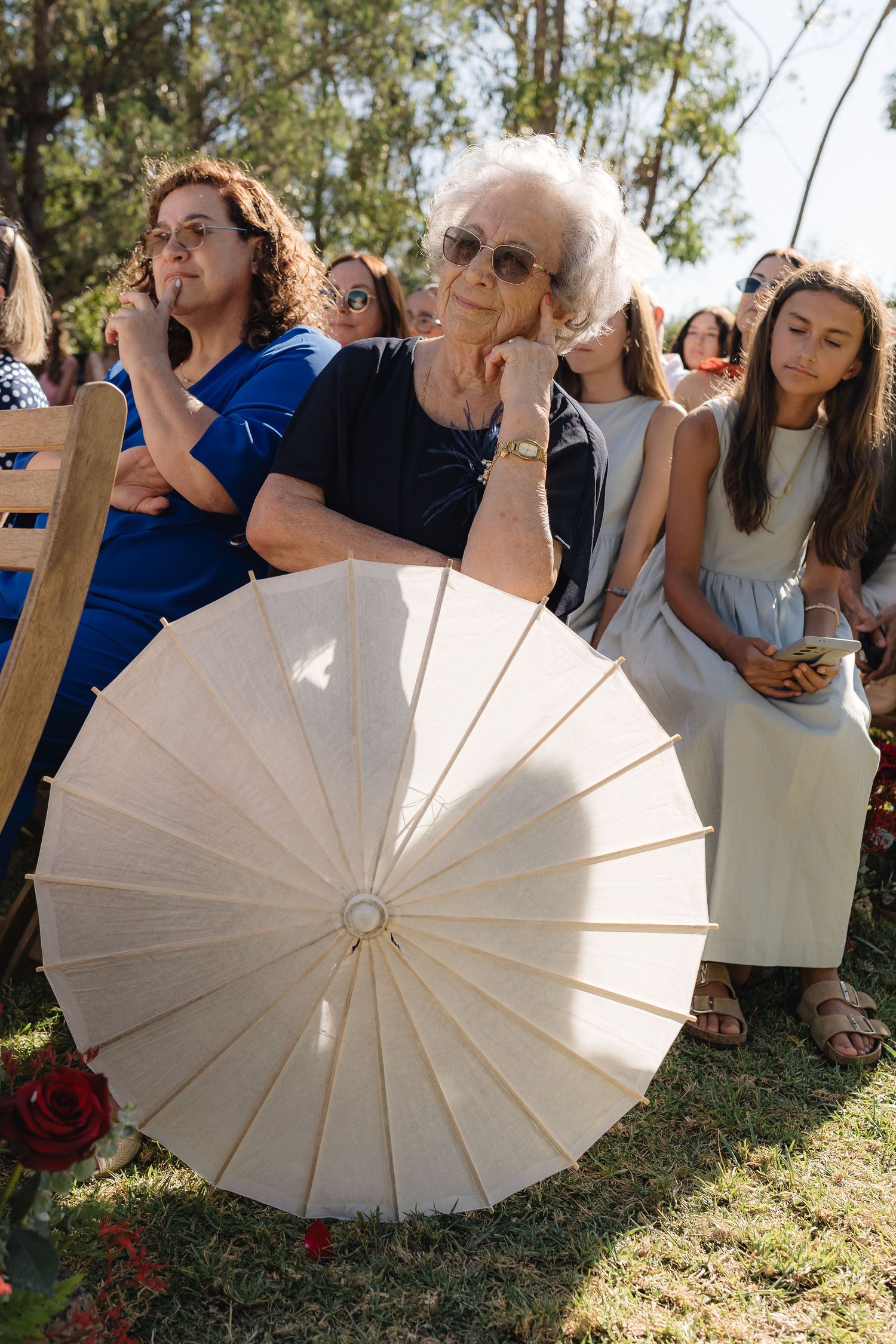 Wedding guests hold white parasols during an outdoor ceremony.
