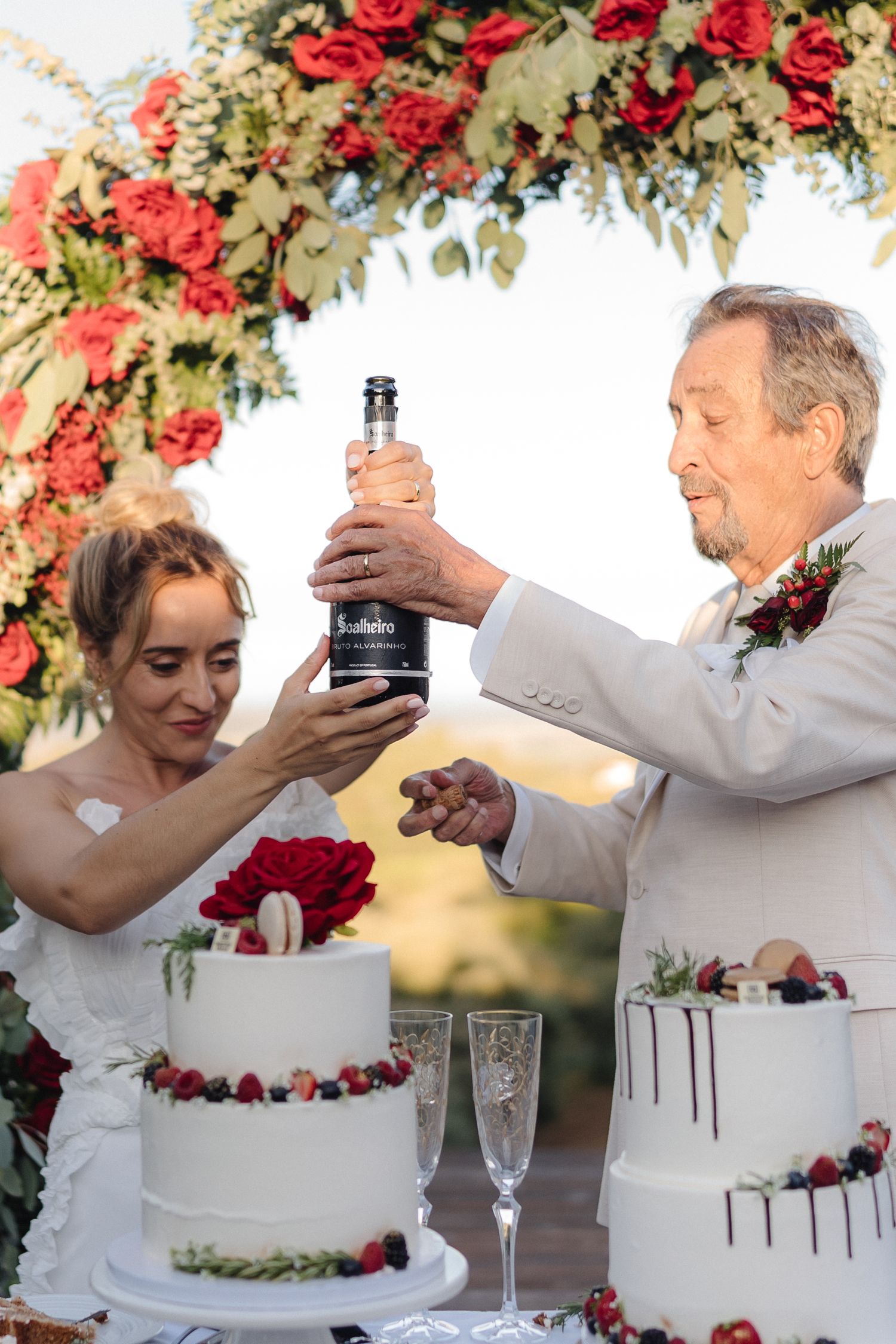 Wedding couple celebrating with cake under a floral arch with red roses.
