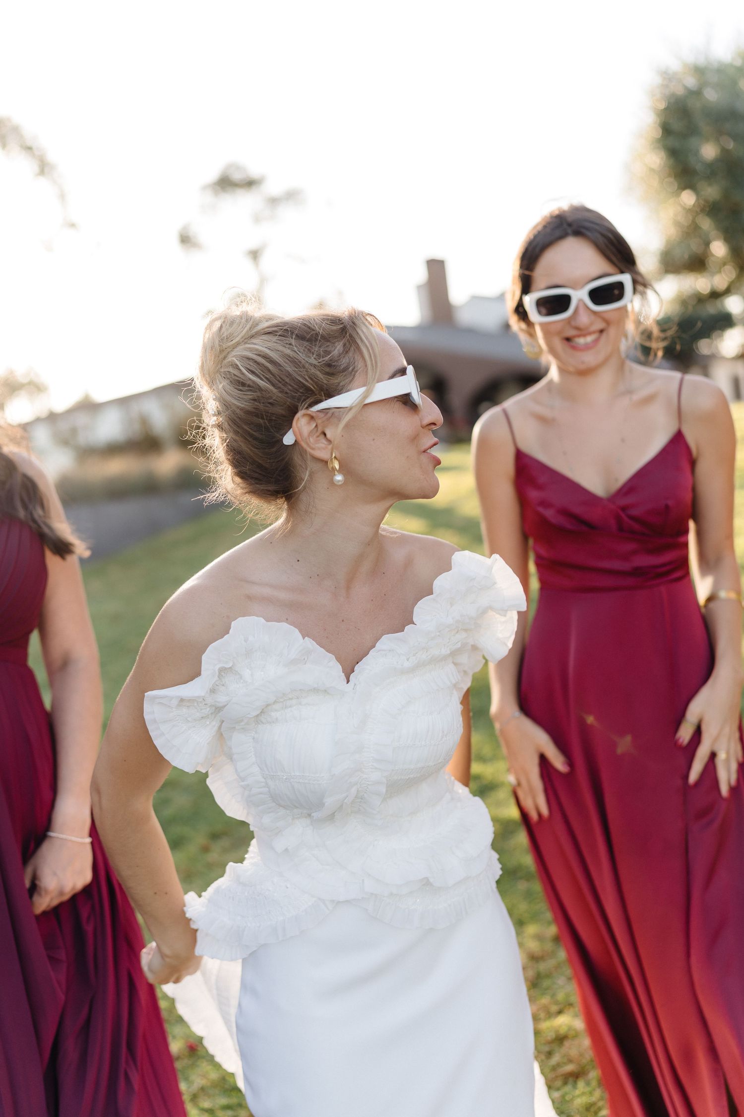 Bridal party in burgundy dresses and white sunglasses dances together on a sunny hillside.