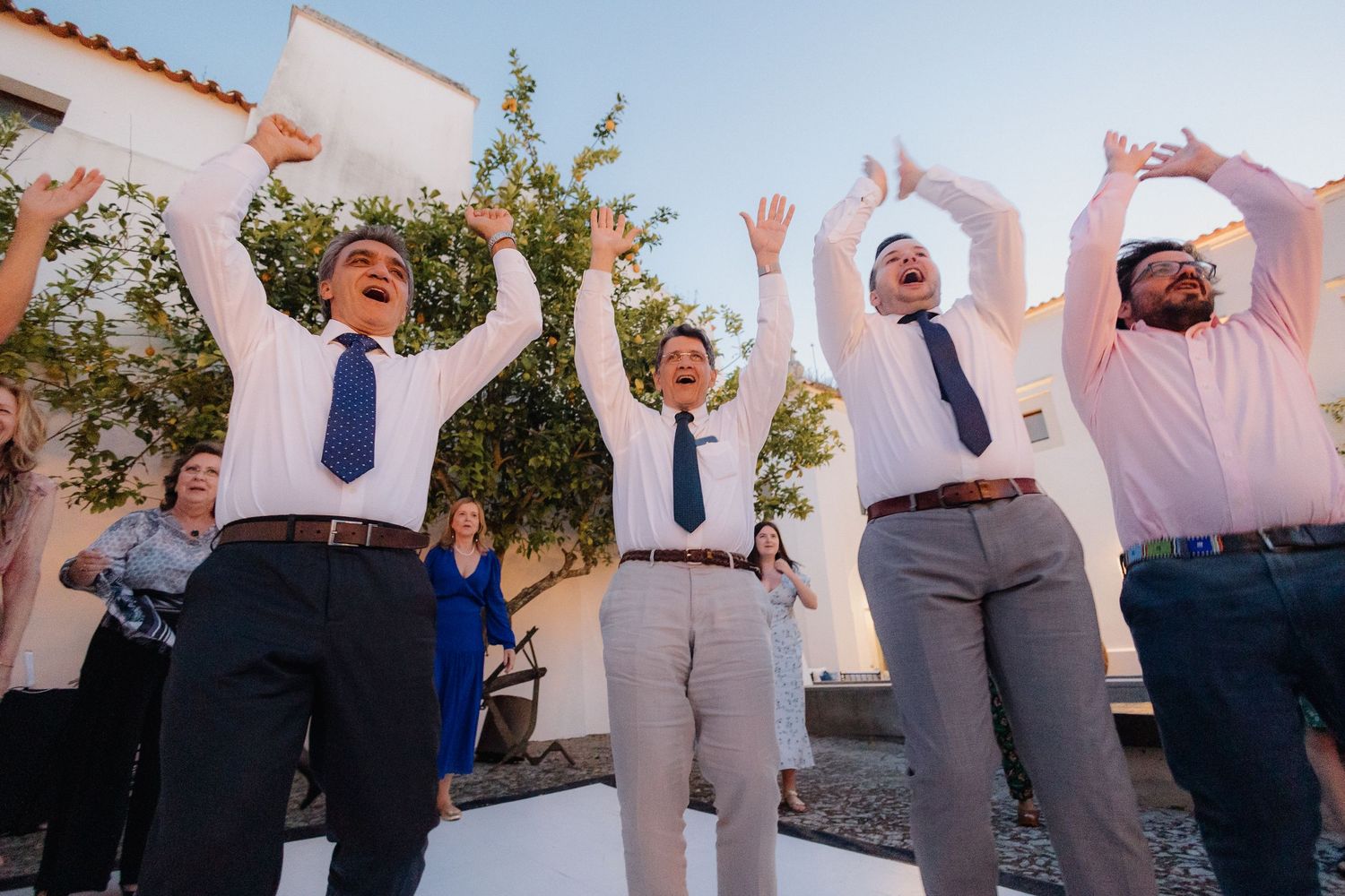 Group of wedding guests jumping and posing for photo against white building.