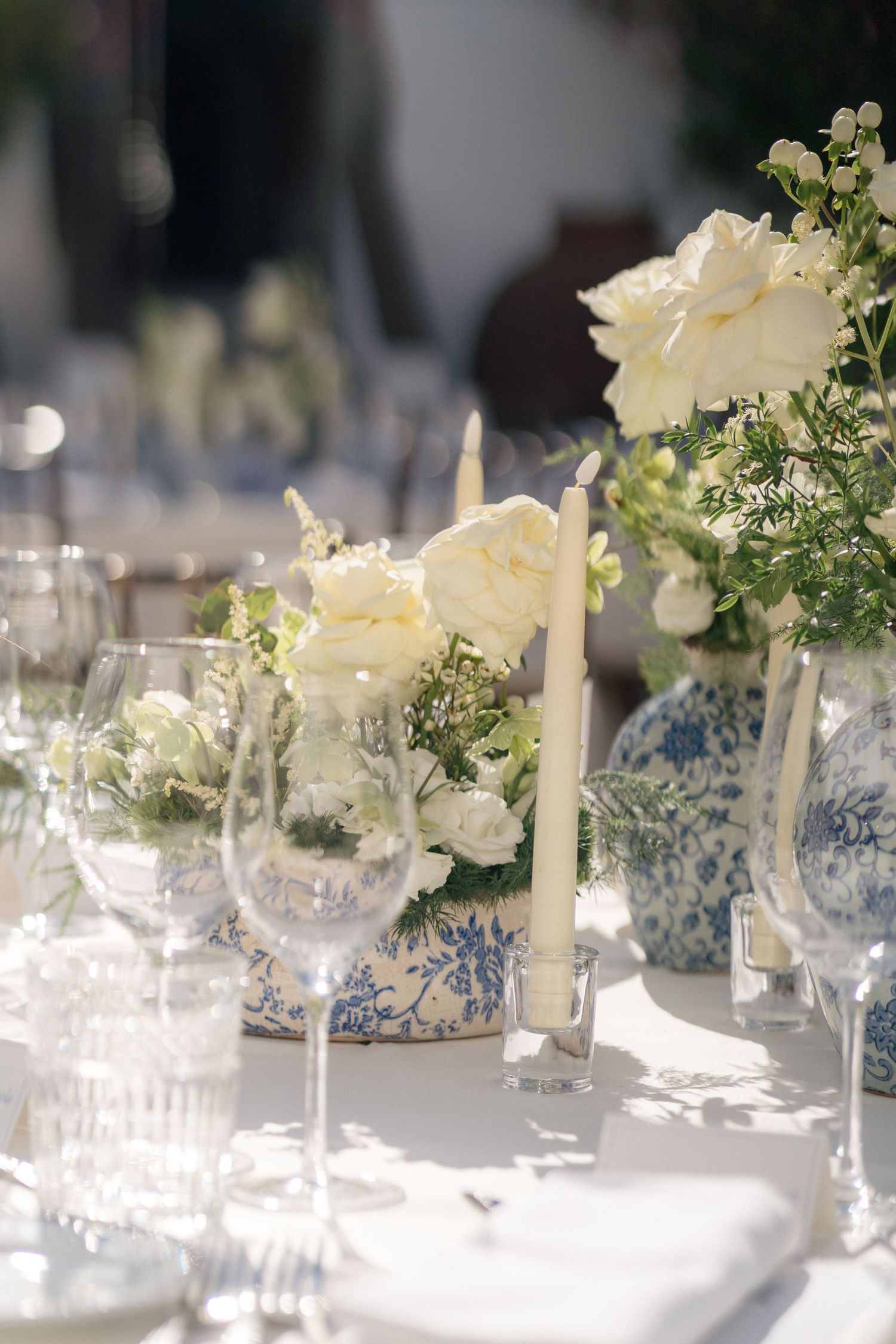 Elegant table centerpieces featuring white roses and blue chinoiserie vases in natural sunlight.