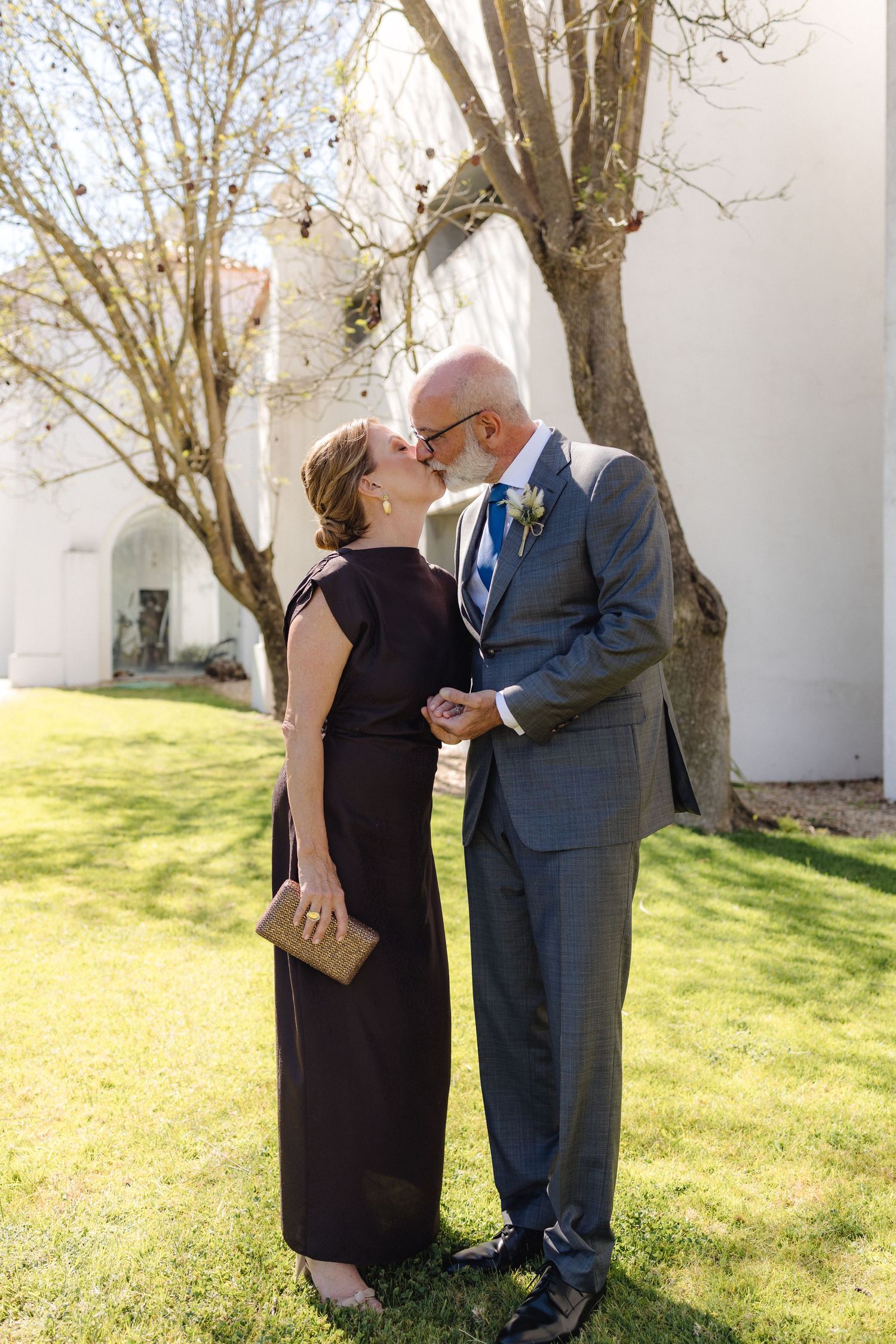 A romantic moment between wedding guests beneath blooming trees outside a white building.