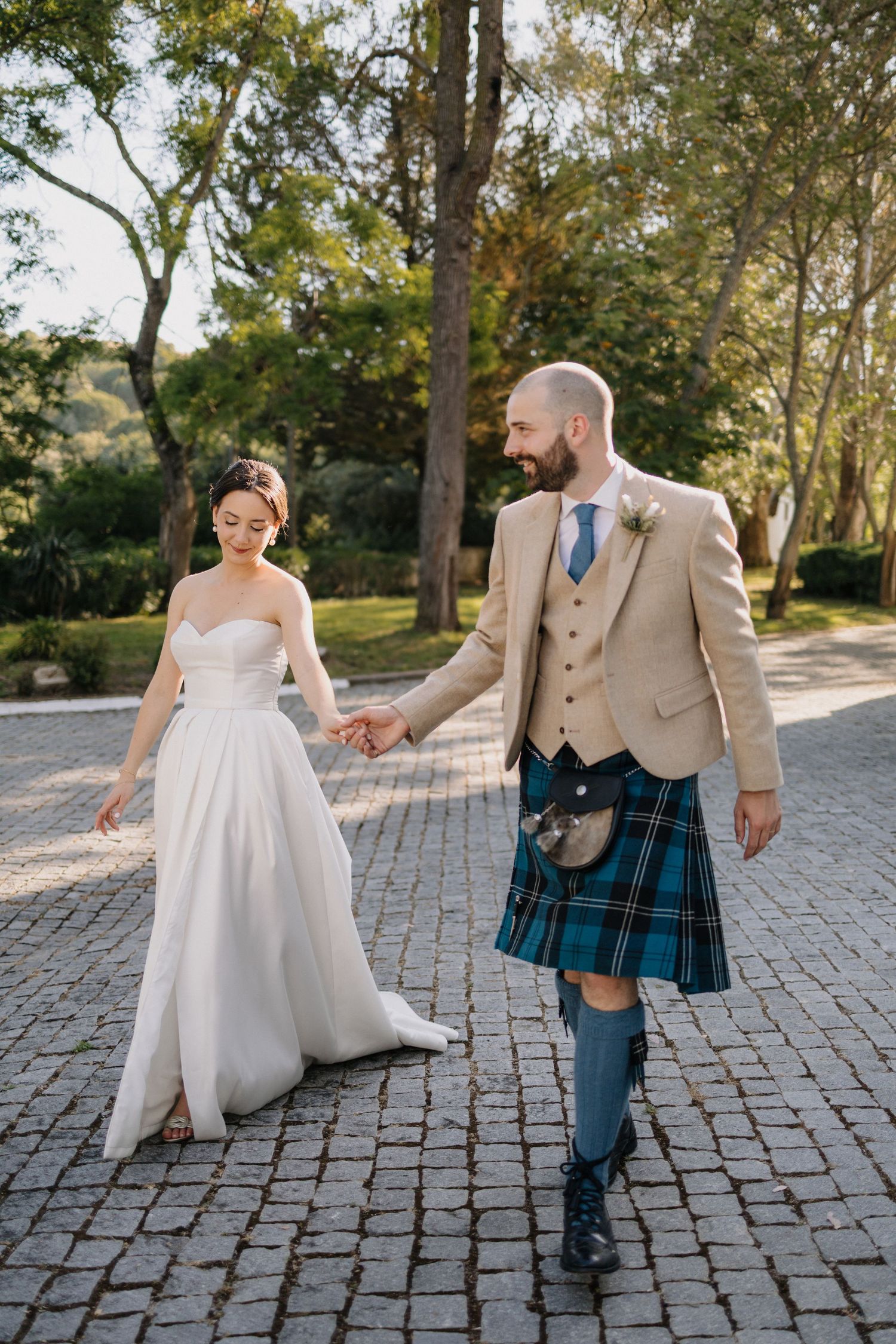 Wedding couple walks playfully on stone path surrounded by trees.