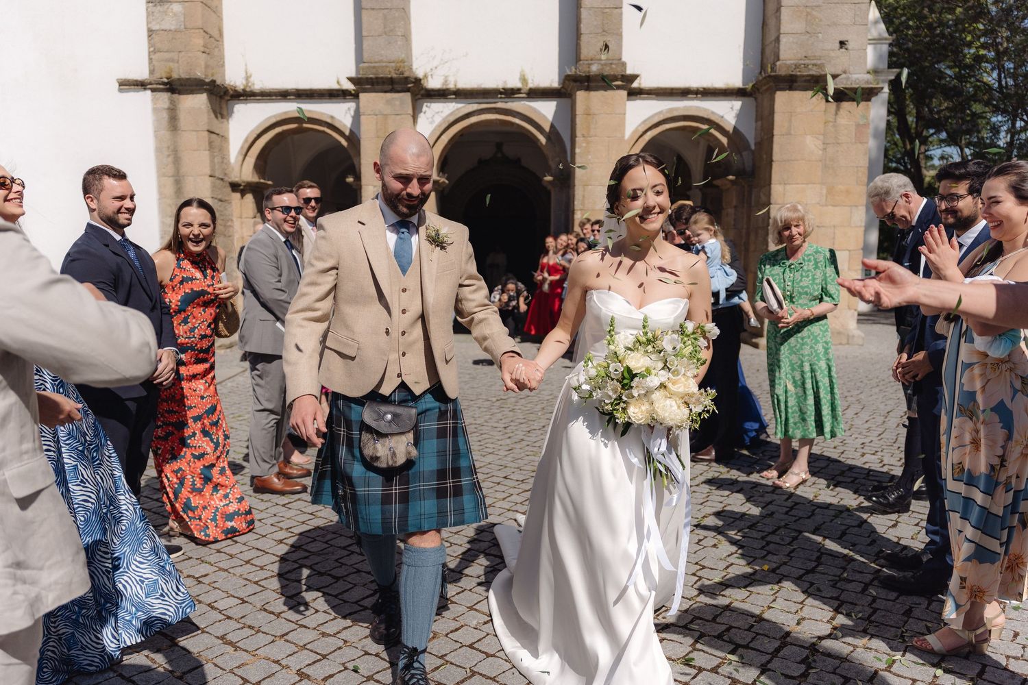 Wedding guests gather outside a stone church building on a sunny day.