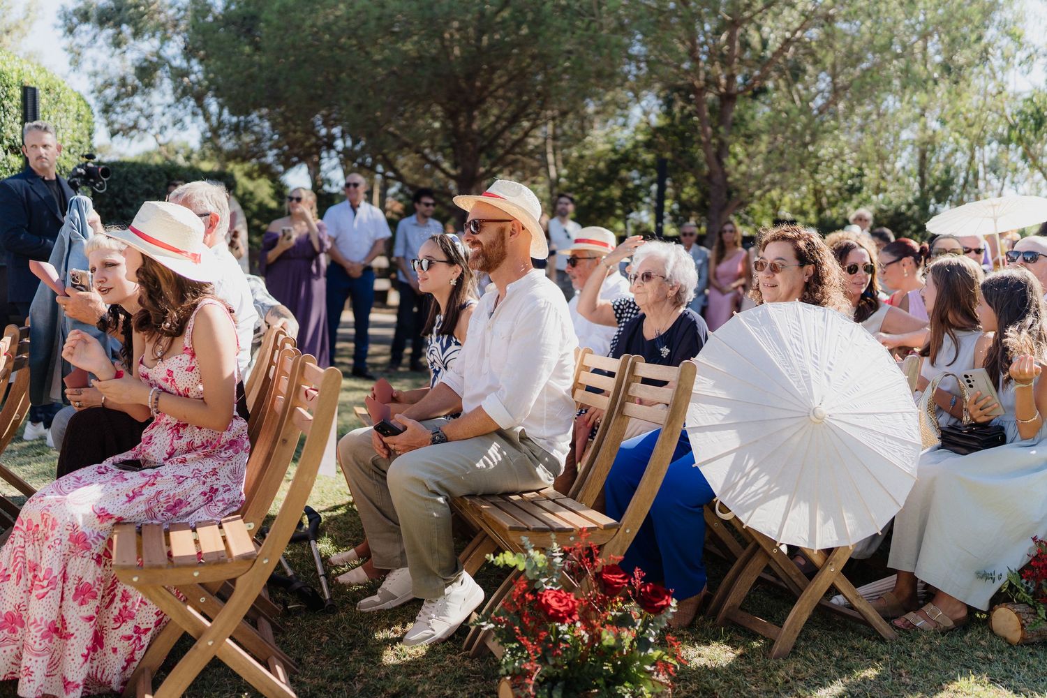 Wedding guests seated in wooden folding chairs under trees at outdoor ceremony venue.