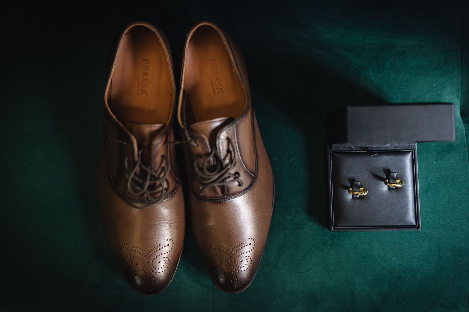 Brown leather dress shoes and ring box arranged on dark green surface.