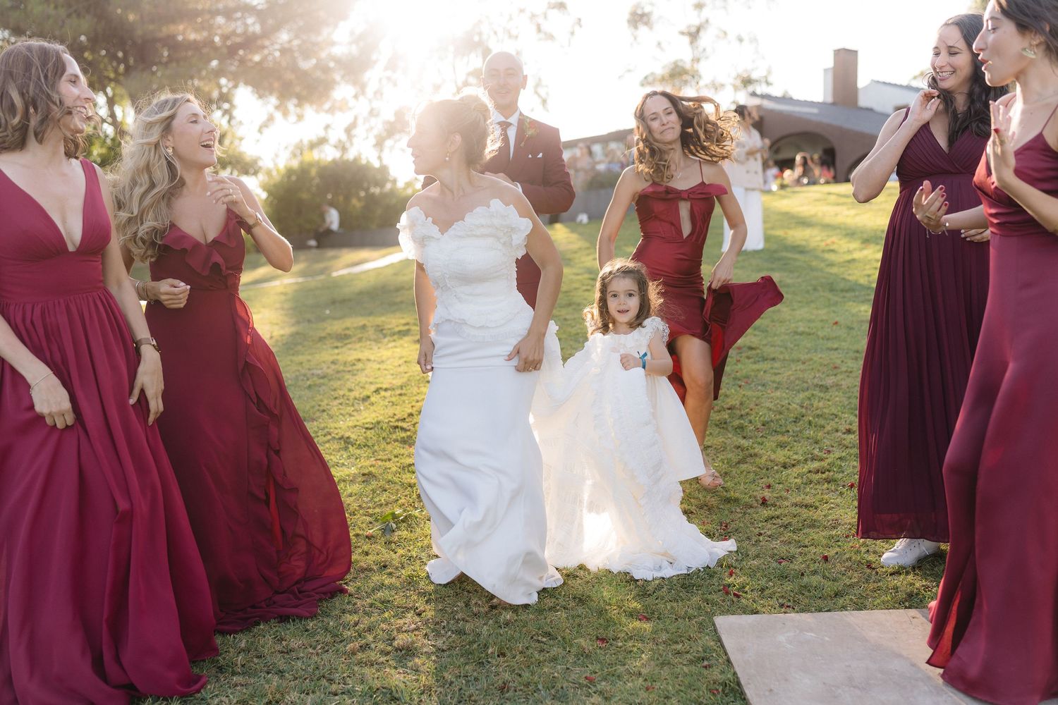 Wedding celebration on grass with burgundy-dressed attendants and white-dressed flower girl.
