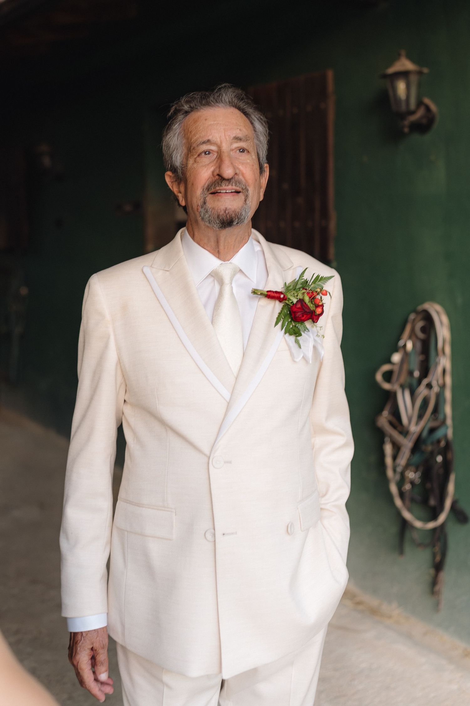 A person in a white suit with a red boutonniere stands in front of an antique wagon wheel.