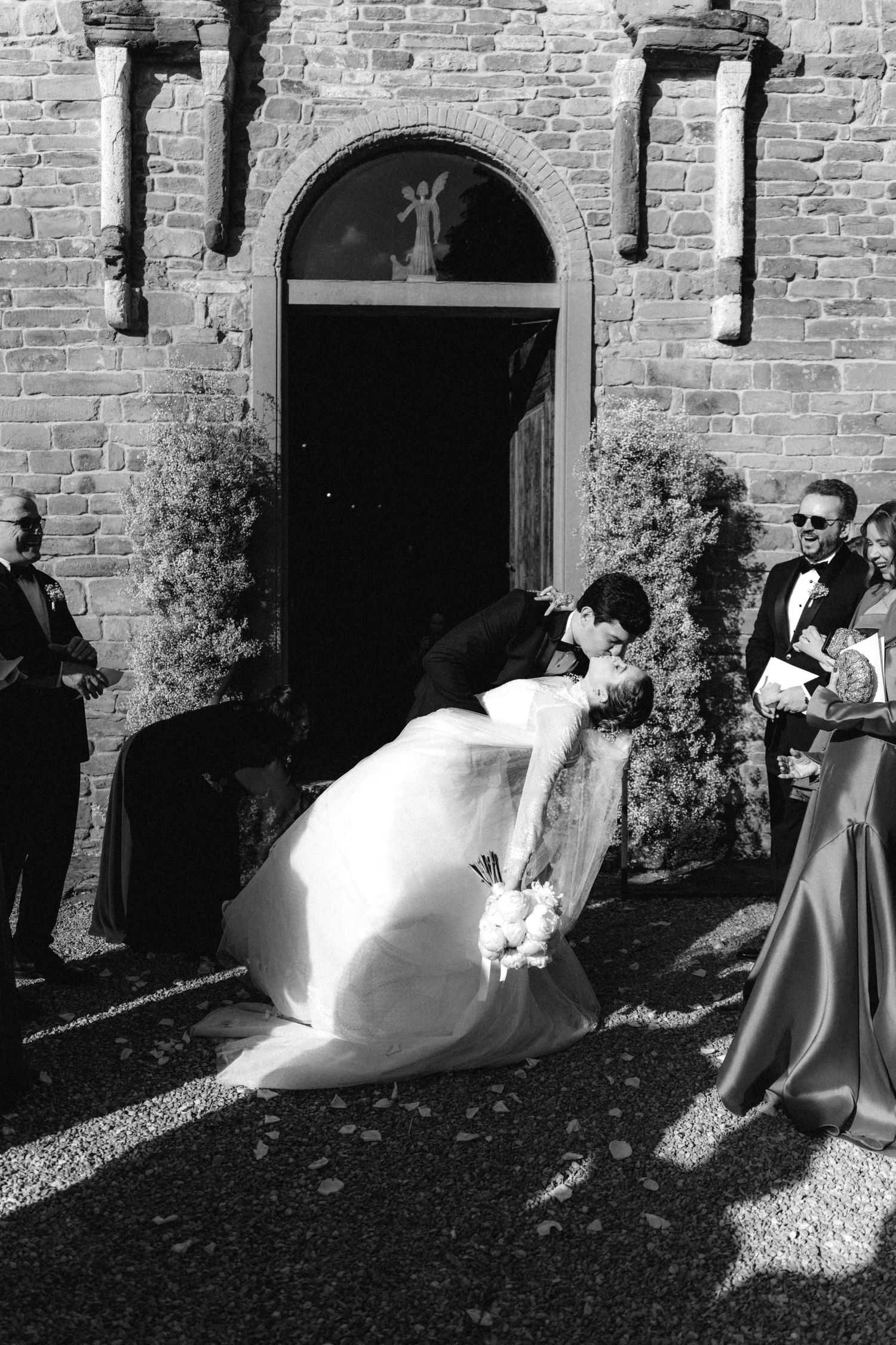 Black and white photo of a groom dipping his bride for a kiss outside a historic stone church after their wedding ceremony.