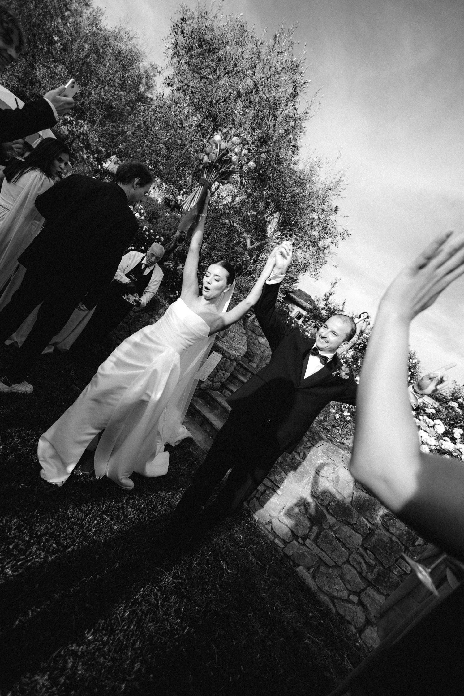 Black and white photo of bride and groom dancing joyfully outdoors at wedding celebration with guests.